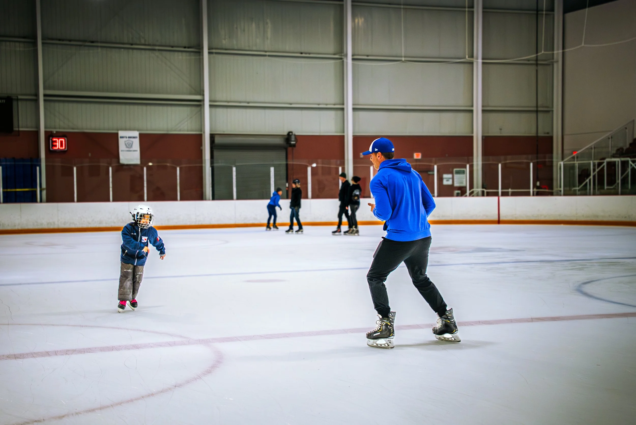 Children and a coach skating on an indoor ice rink with a brick wall and large panels in the background.