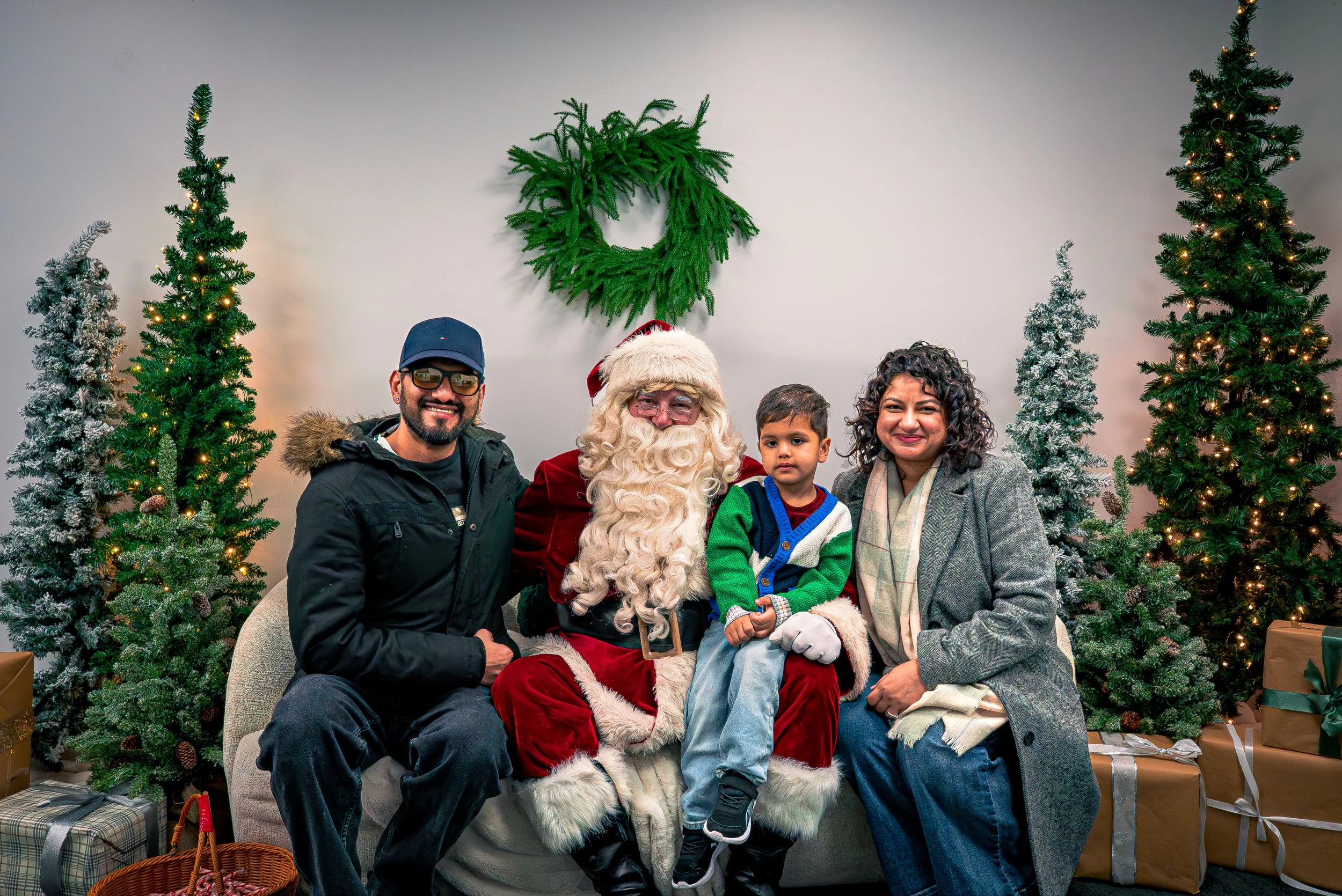 A family of four, including a young boy, sits with Santa Claus in front of decorated Christmas trees. The family is smiling and dressed warmly, with presents and gift baskets on the floor.