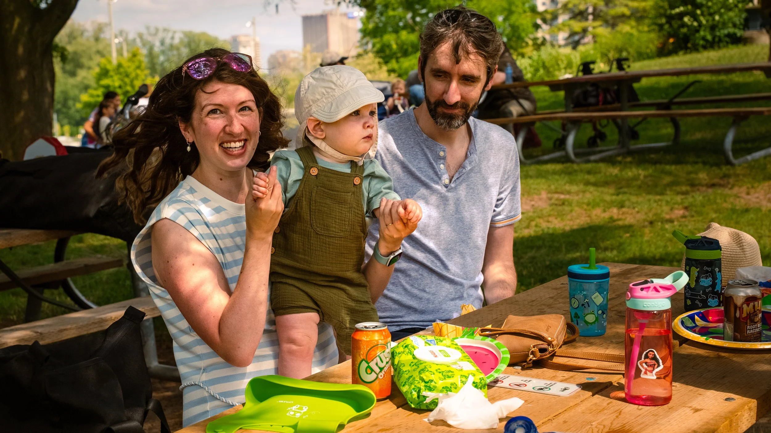 A family of three sitting at a park picnic table, surrounded by colorful cups, snacks, and toys, enjoying a sunny day with trees and a city skyline in the background.