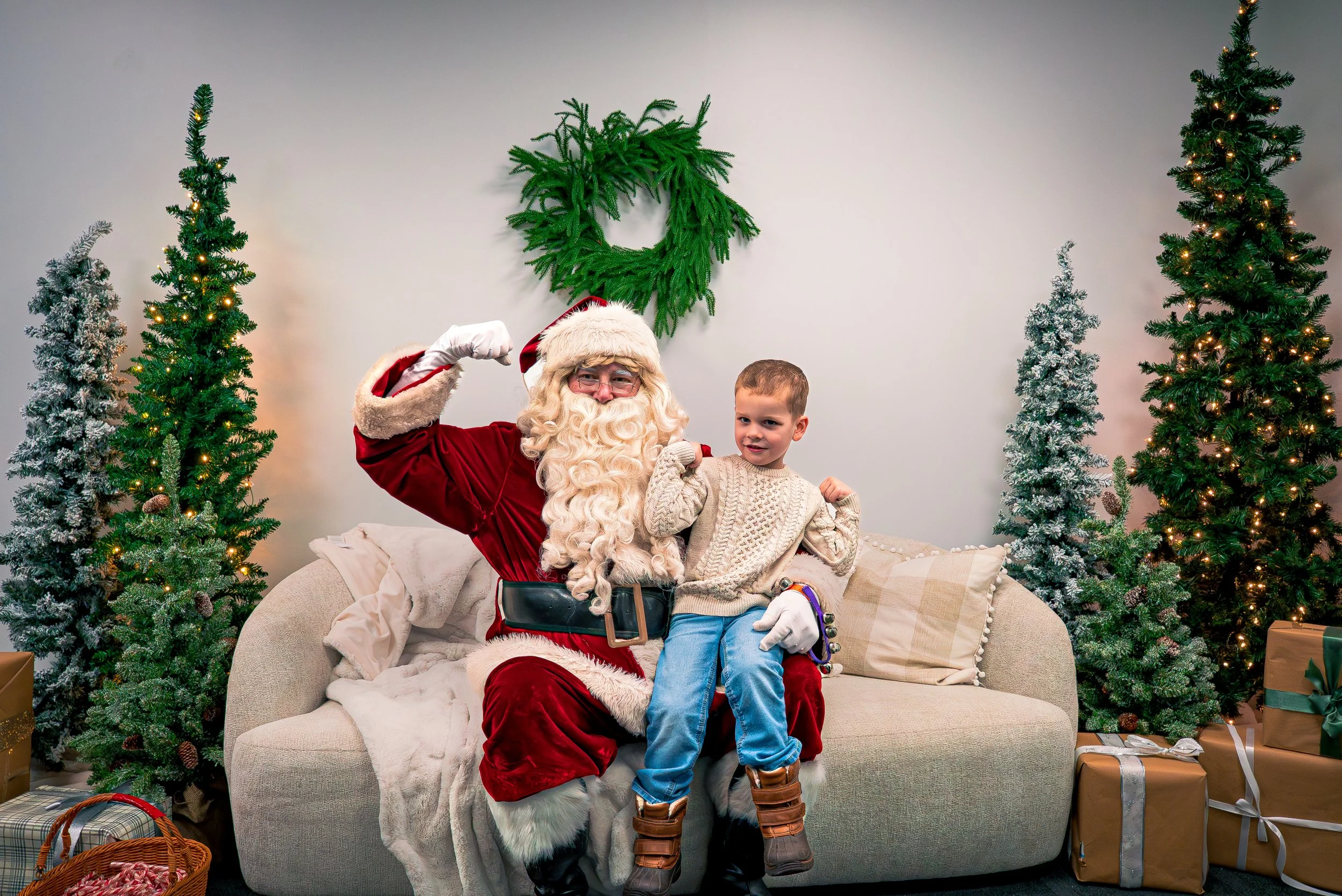 Santa Claus in a red suit and white beard sitting on a beige couch with a young boy, surrounded by decorated Christmas trees and wrapped presents.