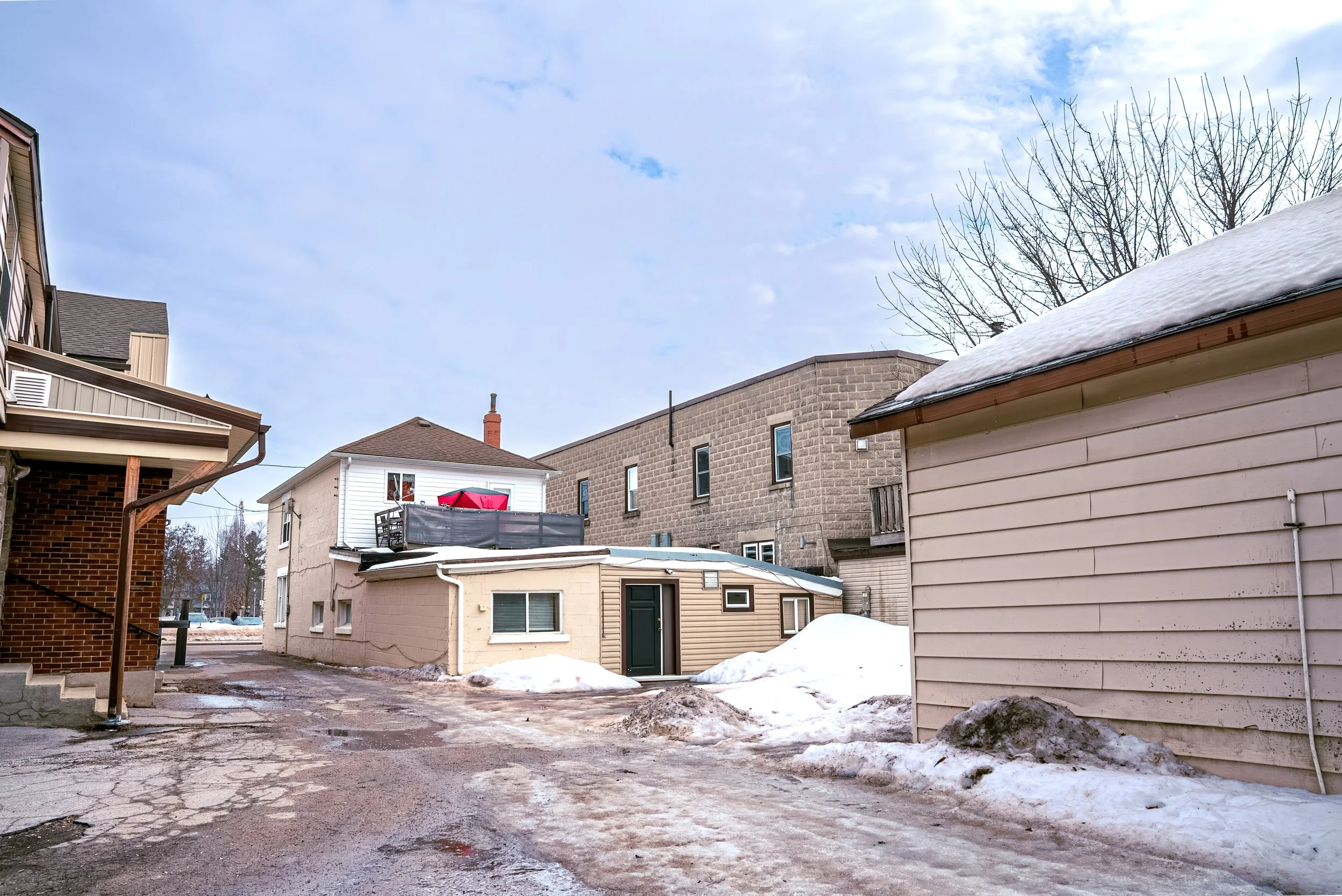 Residential backyard with snow on the ground, partially cleared pavement, and neighboring houses under a partly cloudy sky.