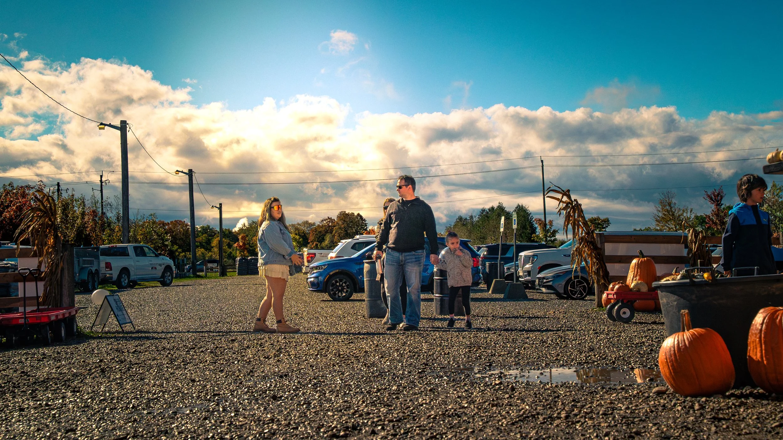People at a fall outdoor market with pumpkins, pumpkins, and farm decorations in a gravel lot, under a partly cloudy sky.