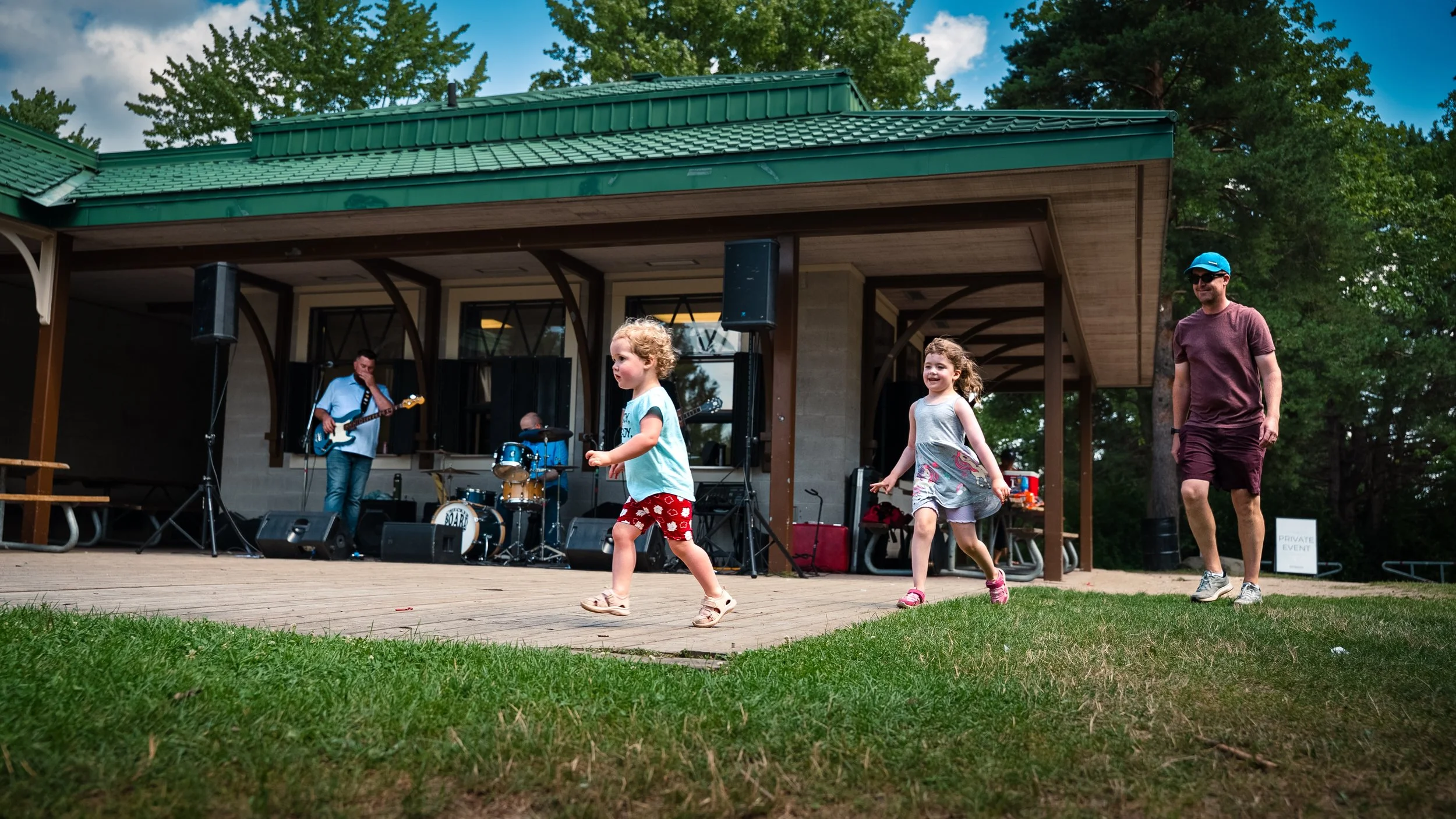 Two young girls dancing and smiling while an adult man watches at an outdoor concert stage with live band playing. Lush green trees surround the scene.