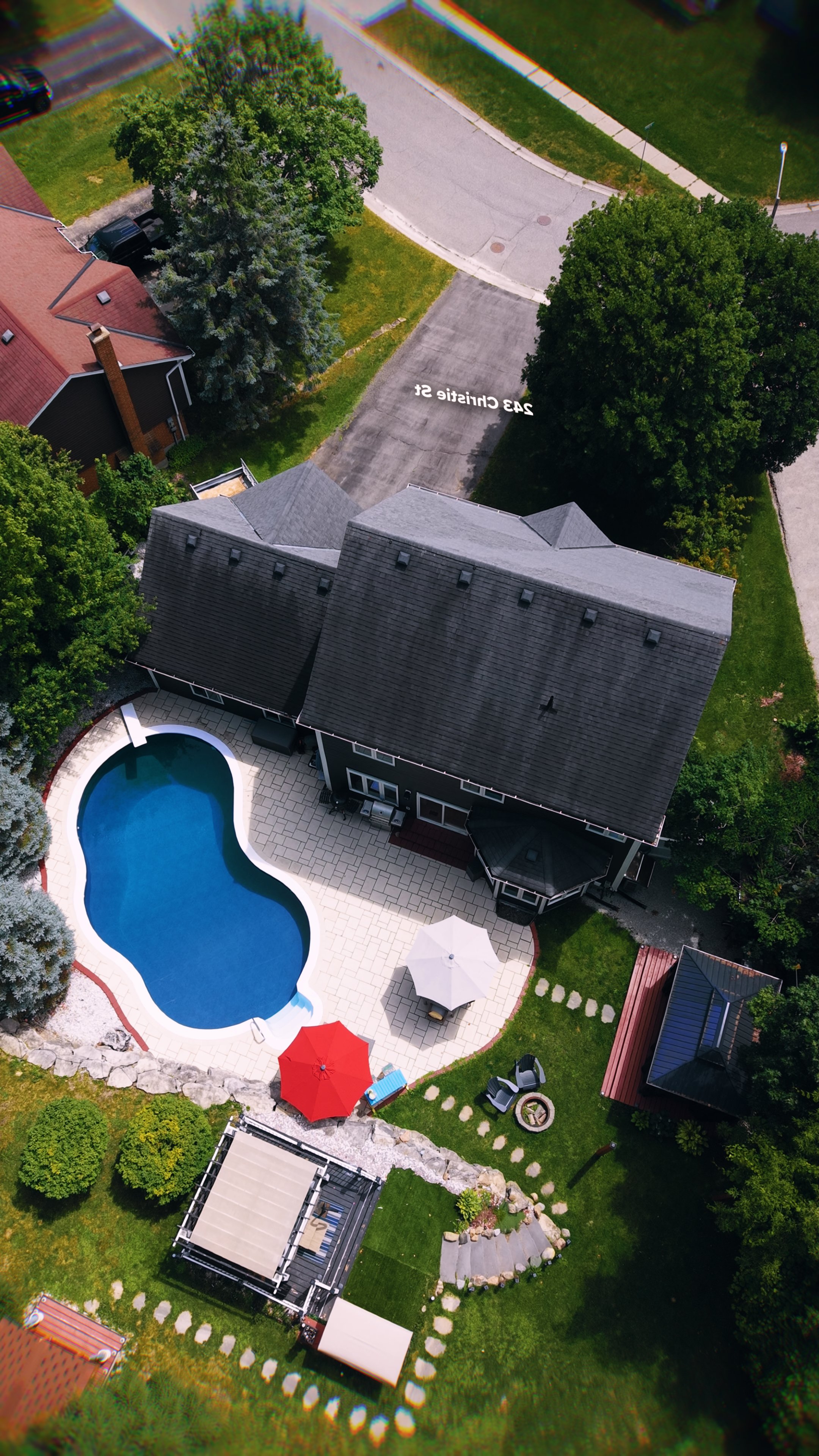Aerial view of a backyard with a kidney-shaped swimming pool, two red umbrellas, patio furniture, green lawn, trees, and neighboring houses.