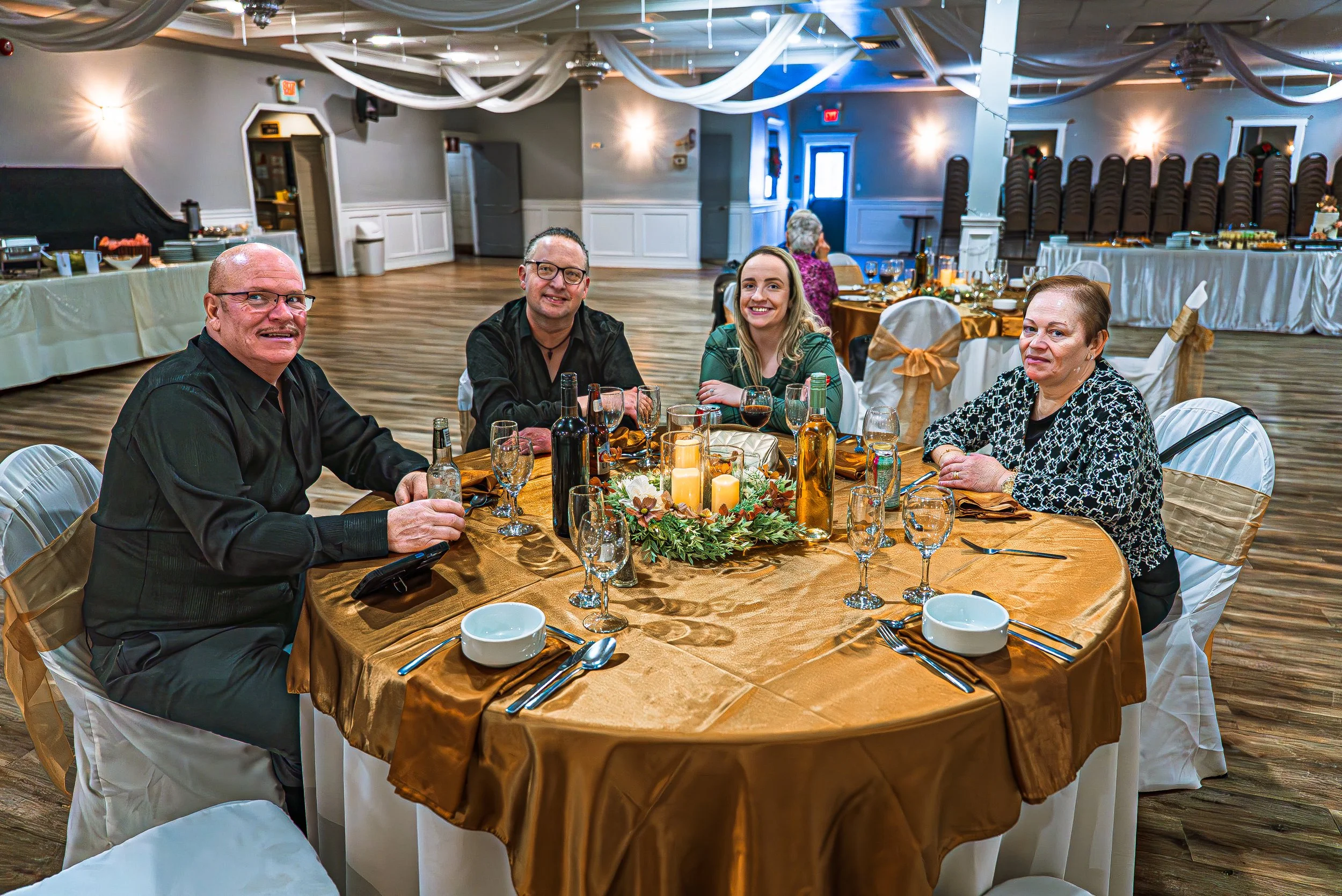 Four people sitting at a round banquet table decorated with a gold tablecloth, candles, and floral centerpiece in a decorated event hall with draped ceiling and chairs with gold bows.