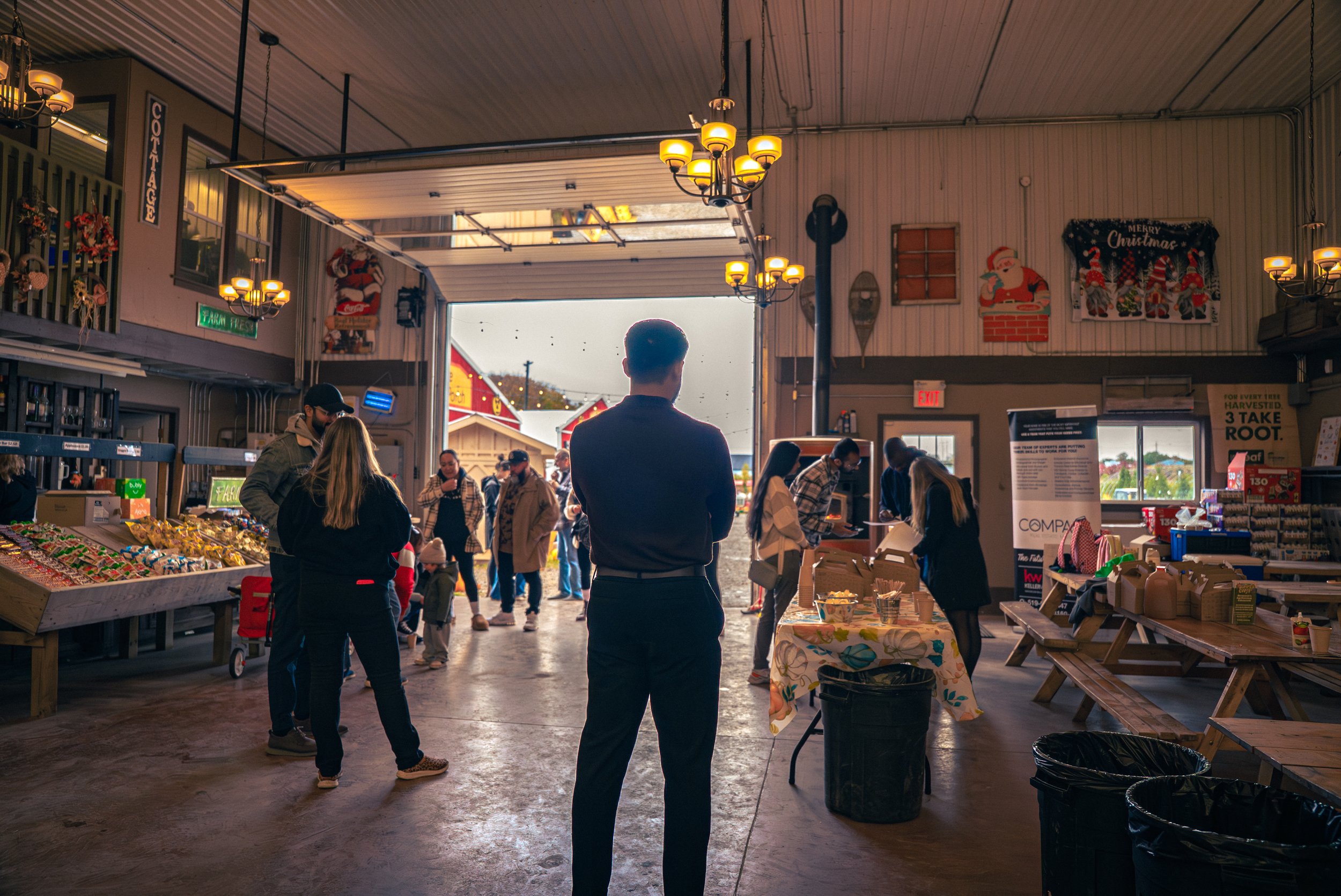 People inside a rustic indoor market or farm shop with Christmas decorations, selling produce and baked goods, with some people browsing and shopping.