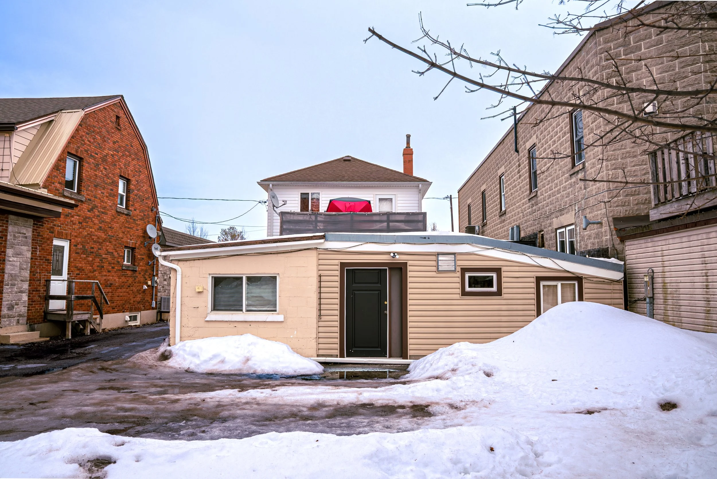 Backyard scene with a small beige building, snow on the ground, and larger brick and stone buildings in the background. Overcast sky.
