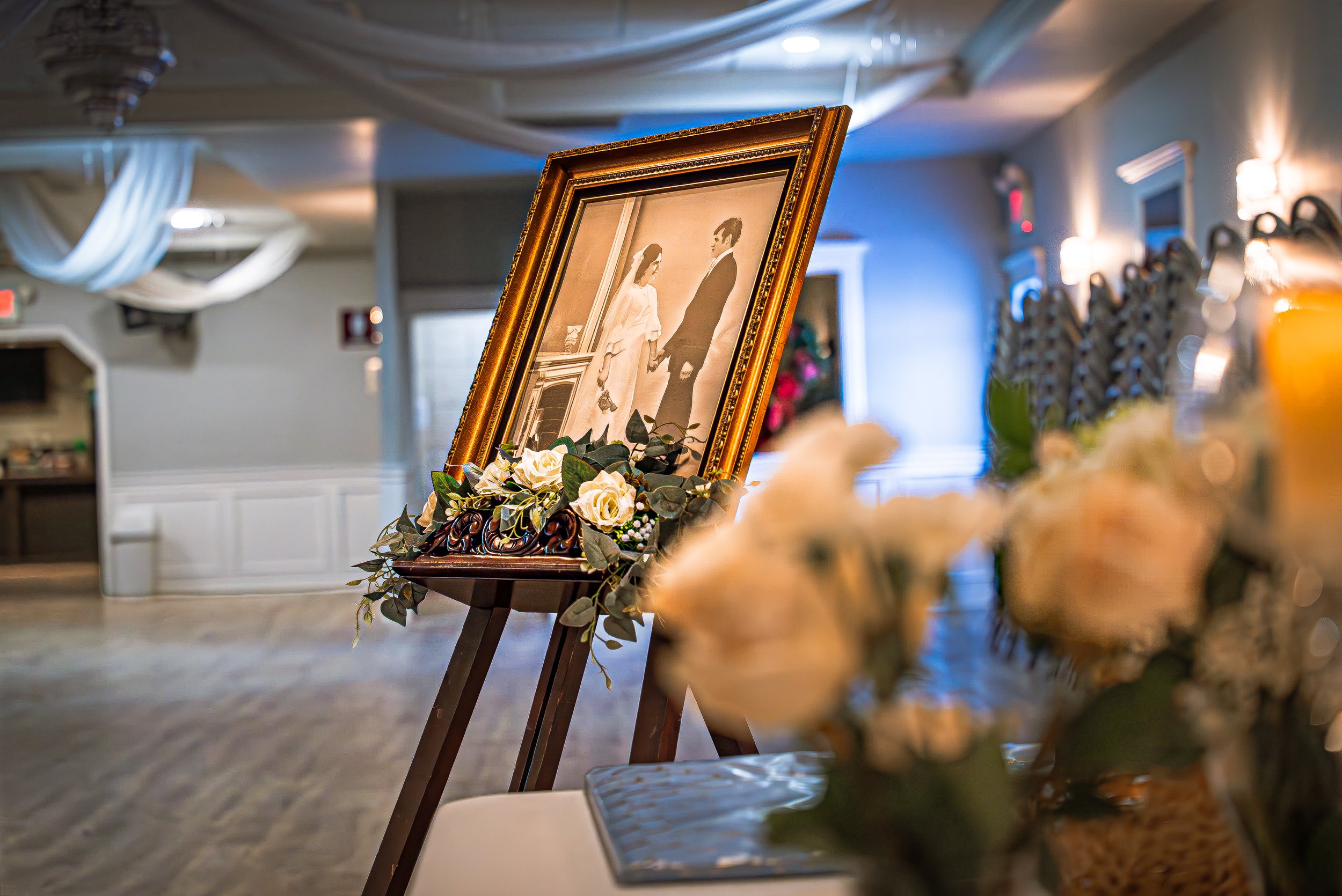 A framed photo of a bride and groom holding hands, displayed on an easel surrounded by flowers, in a reception hall.