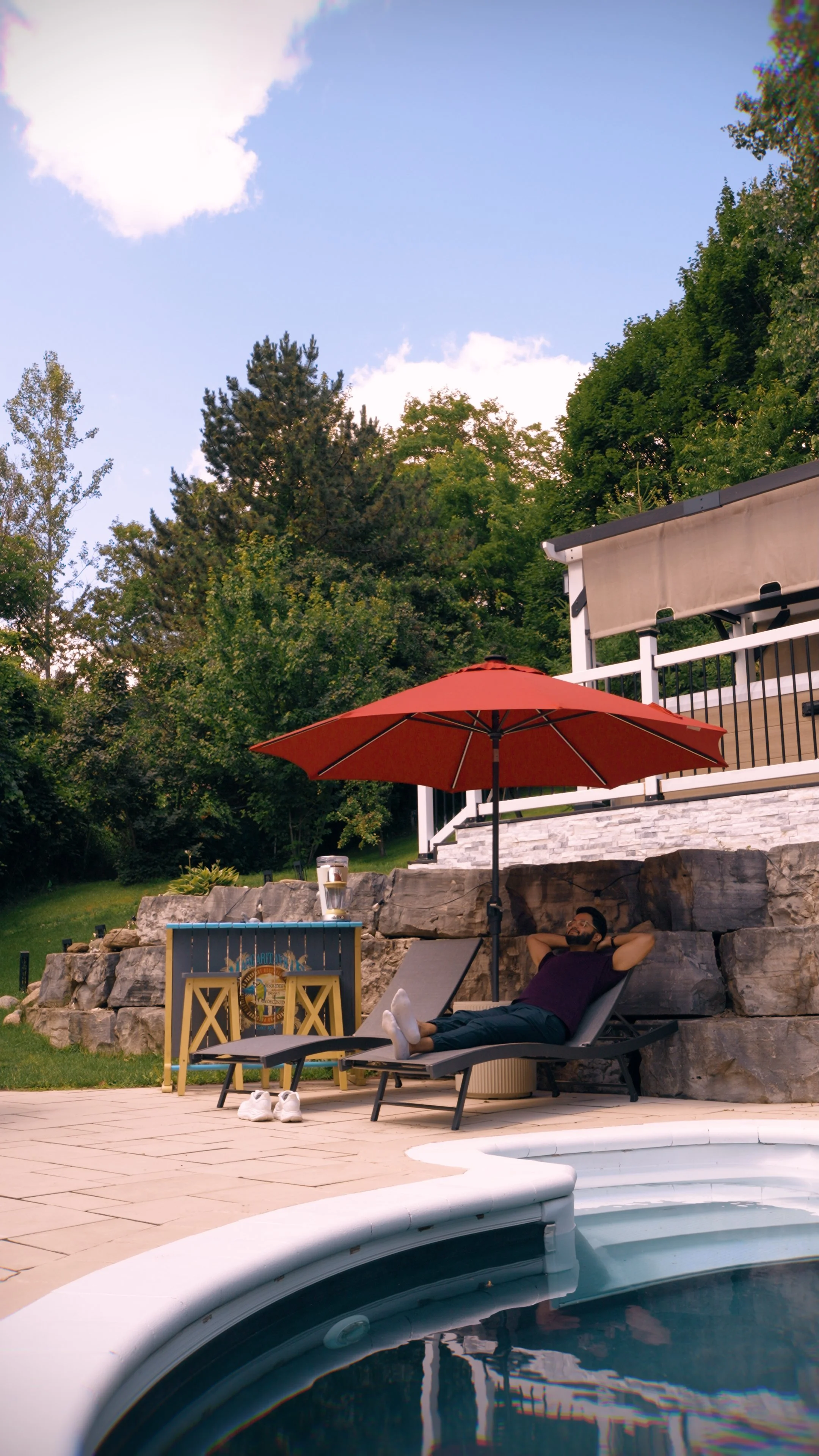 A man relaxing on a lounge chair beside a swimming pool, with a red umbrella shading him. There are large rocks behind him and greenery, including trees and bushes, in the background.