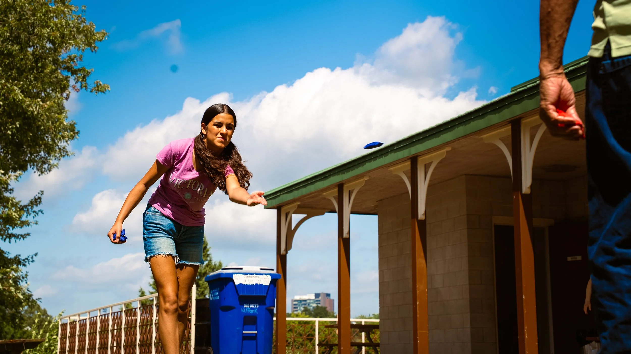 A young woman with long brown hair, wearing a pink shirt and denim shorts, is throwing a blue disc during a game of disc golf outdoors on a sunny day. There are trees and a building with a green roof in the background, and a person partially visible 