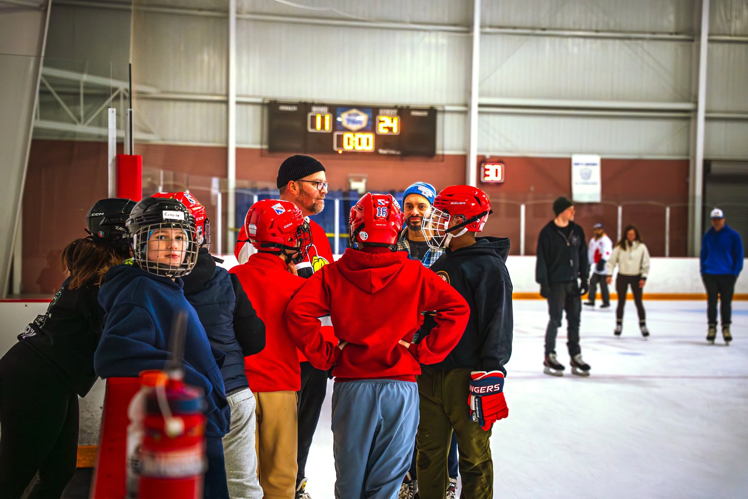 A group of hockey players and coaches gathered on the ice rink during a game or practice, with a scoreboard in the background showing the score as 11-24 and the game time at 0:00.