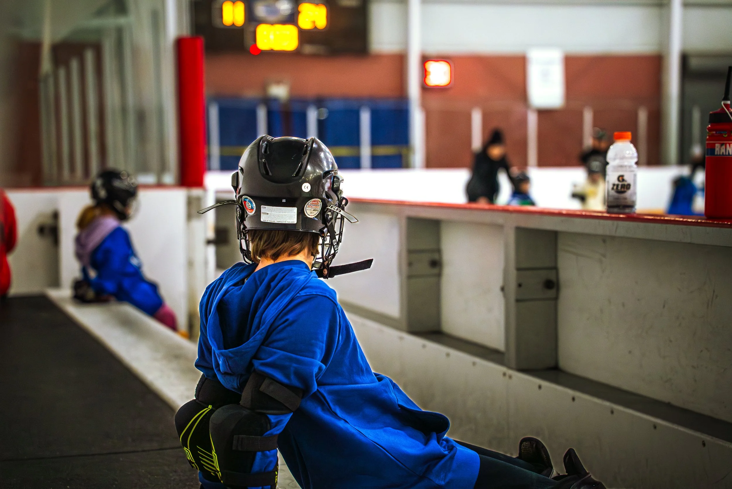 A young child wearing a black hockey helmet and blue hockey gear sits on the ice in a hockey rink, with other children and a coach in the background.