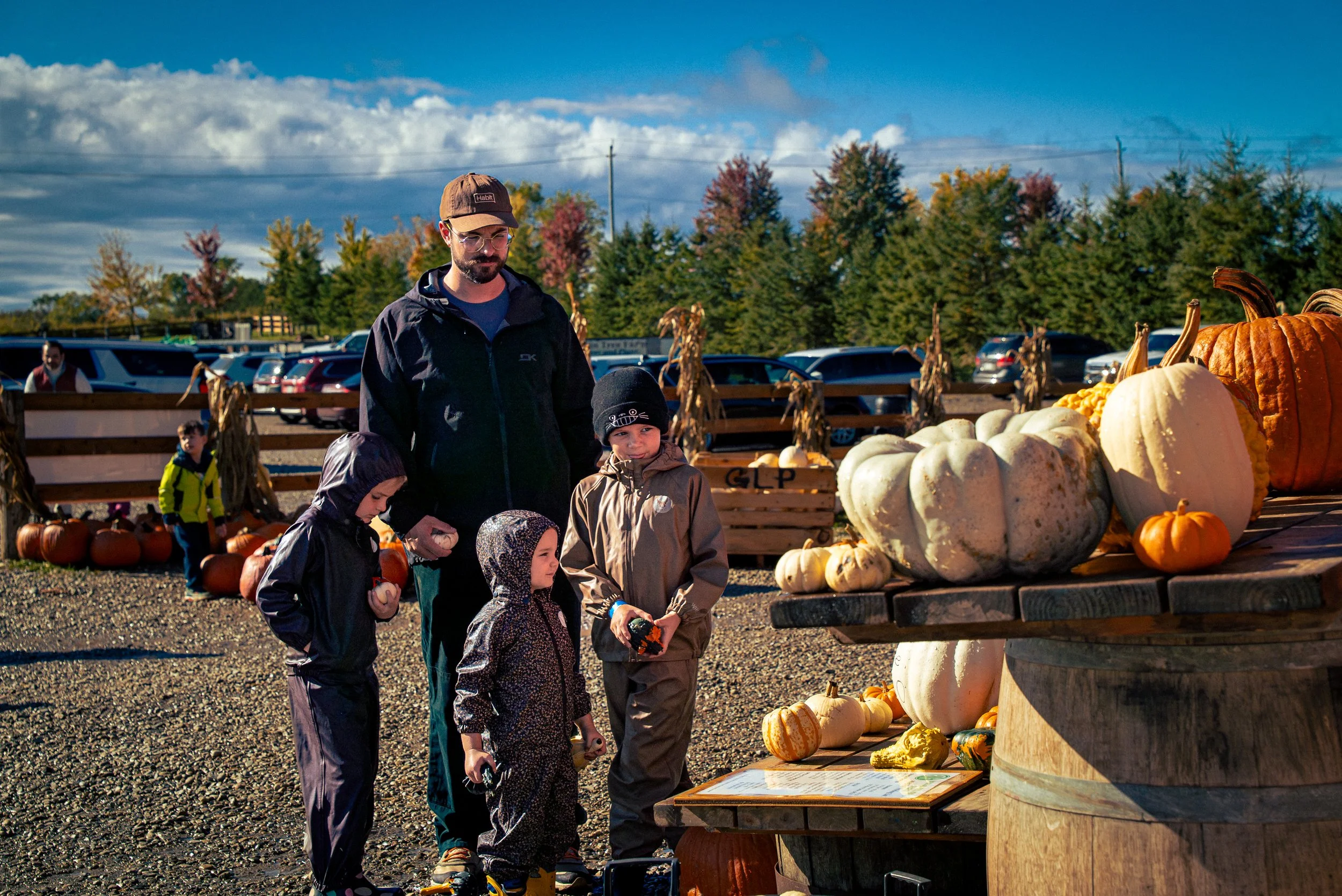 People, including children, at a pumpkin stand outdoors, with various pumpkins on display and trees with fall foliage in the background.