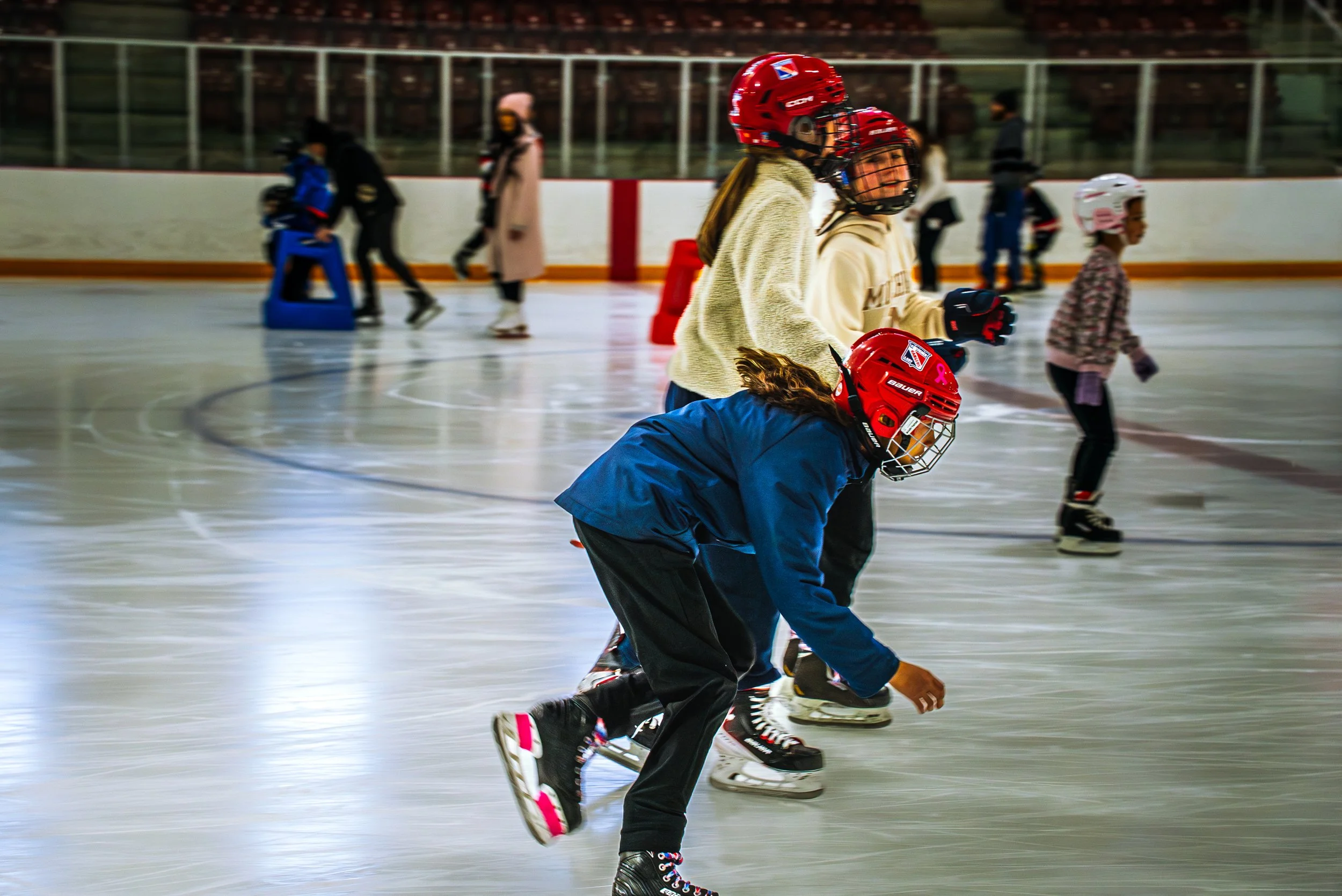 Children ice skating on an indoor rink, some wearing red helmets and skating gear, others using support aids, with a background of a barrier and seating area.