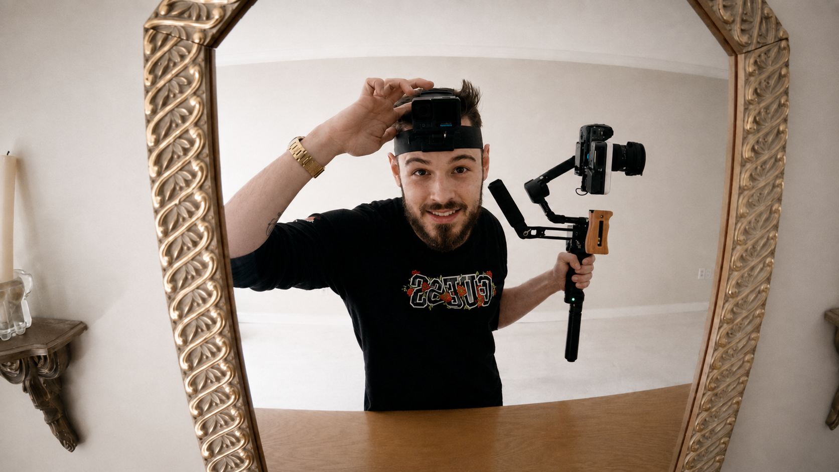 A man taking a selfie in a mirror with a camera mounted on a stabilizer. He is wearing a black shirt with colorful text and a gold wristwatch.