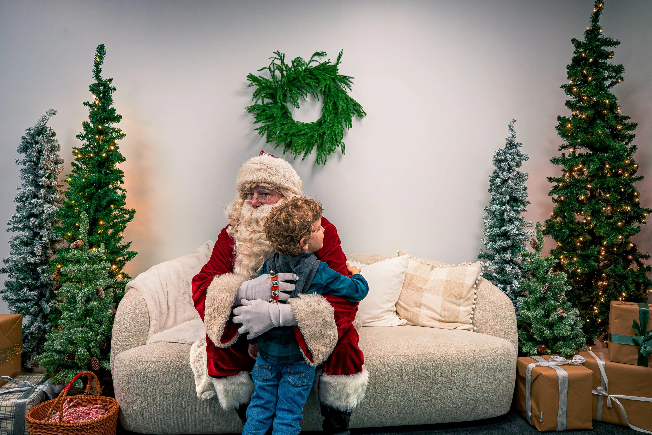 Santa Claus in a red suit with white trim, holding a young boy in a blue shirt, seated on a cream-colored sofa in a Christmas decorated room with Christmas trees, wrapped presents, and a green wreath on the wall.