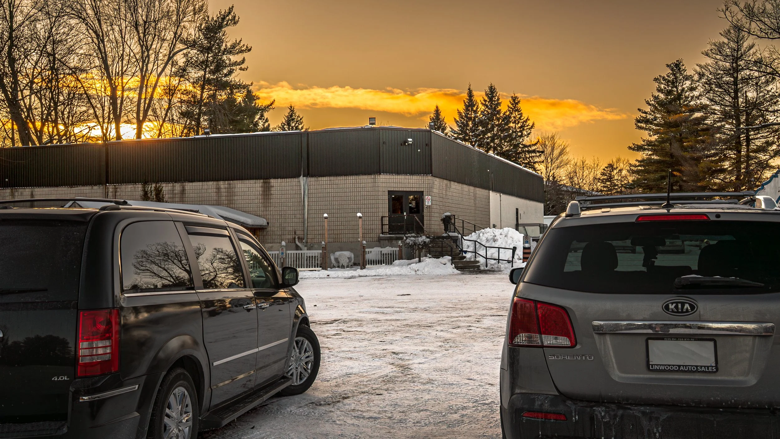Parking lot with two cars in the foreground, snow on the ground, a building in the background, and trees with a sunset sky.