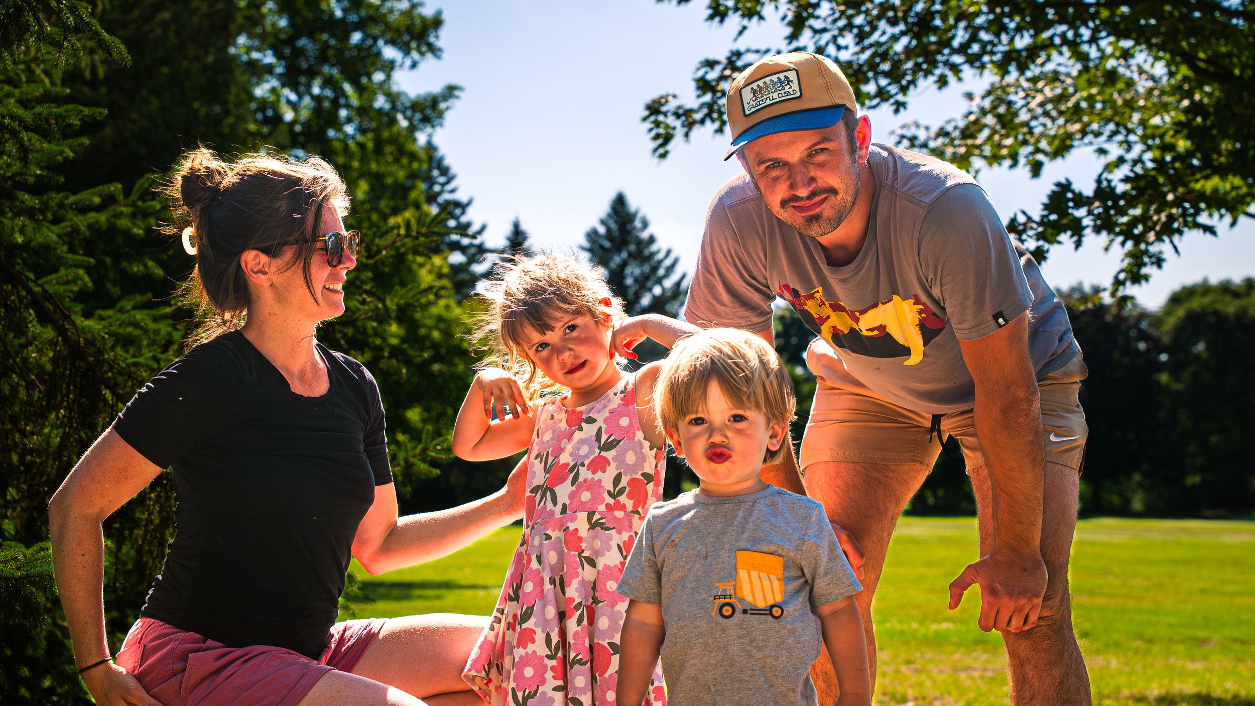 A family of five outdoors on a sunny day, including a woman, a man, and three children, surrounded by trees and greenery.
