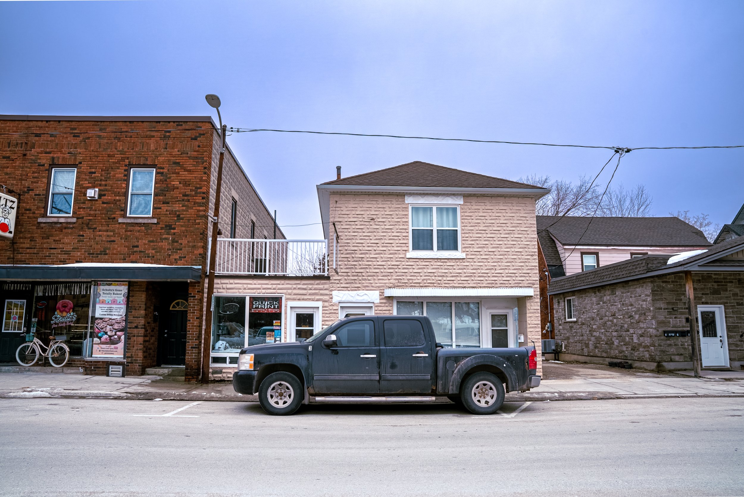 A black pickup truck parked in front of a row of brick and light-colored buildings on a city street under a clear blue sky.