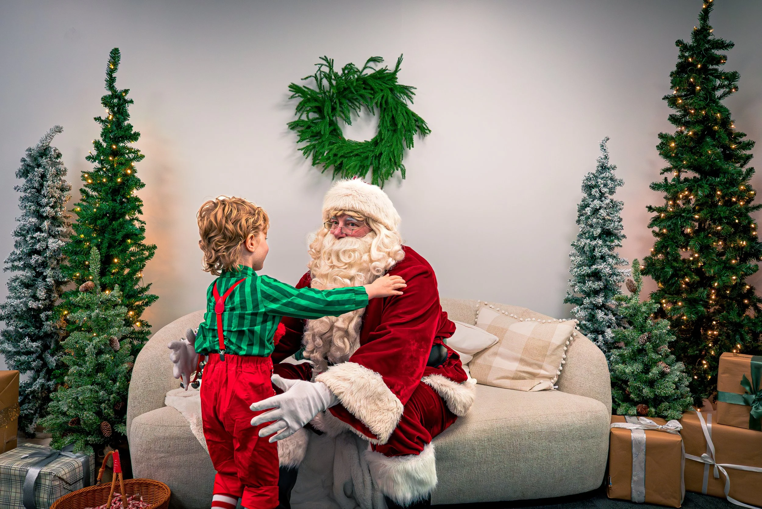 A young boy hugging Santa Claus who is sitting on a beige couch surrounded by decorated Christmas trees and wrapped presents.