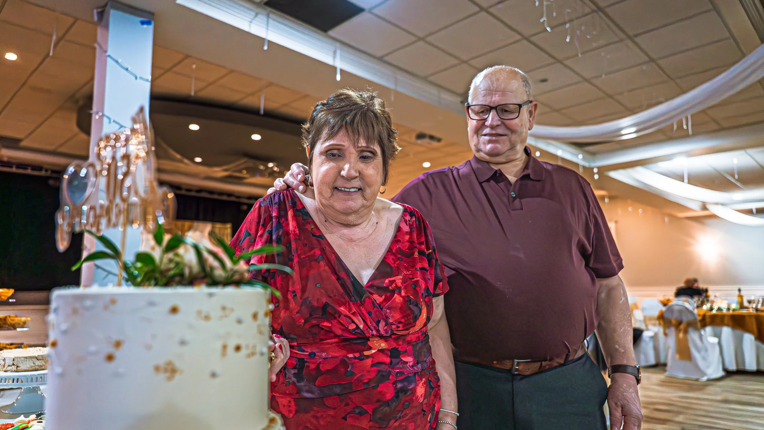 An elderly woman in a red and black floral dress and an elderly man in a maroon shirt and glasses stand together at a celebration, looking at a cake with a crystal decoration and flowers in the foreground, in a decorated banquet hall.
