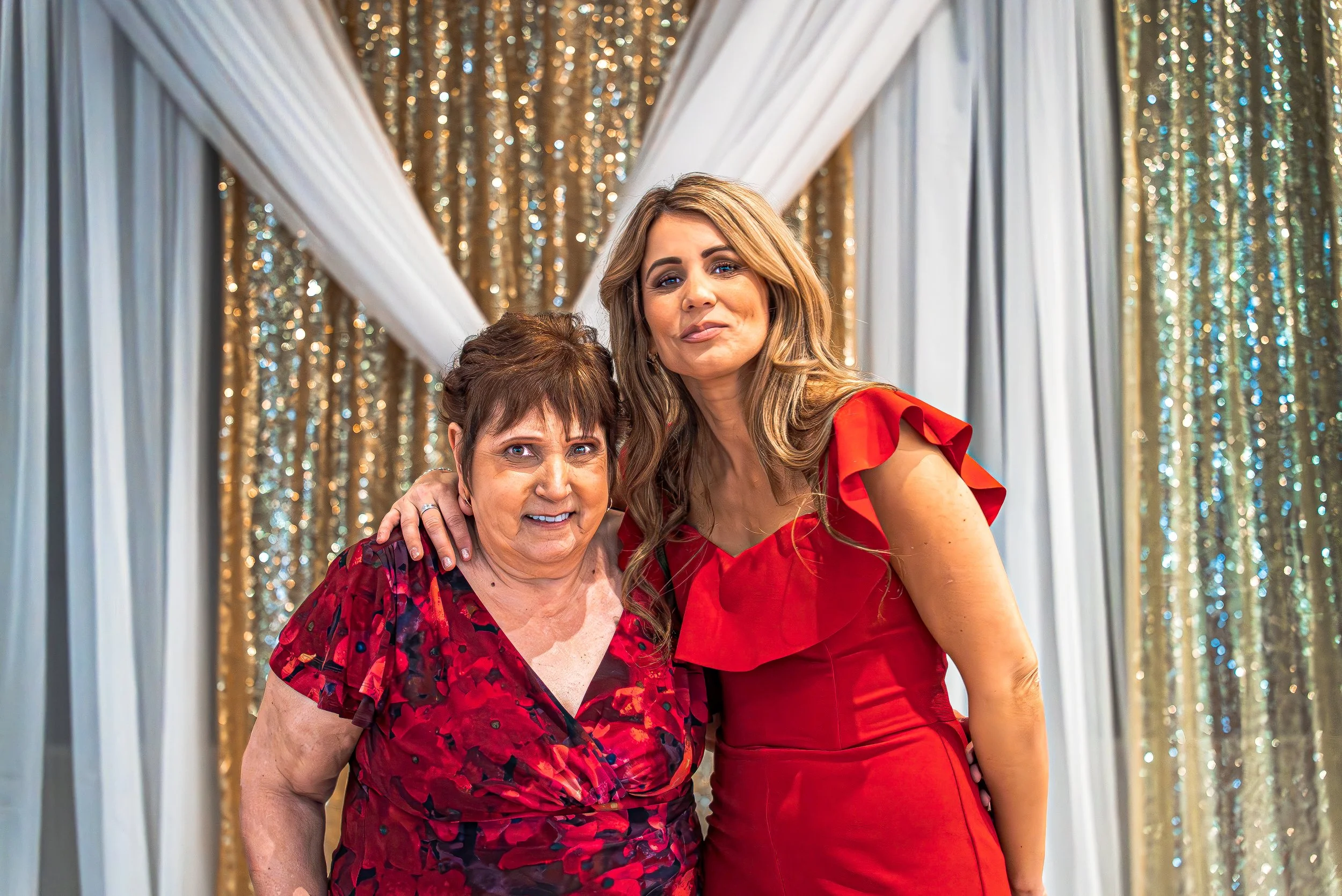 Two women, one older and one younger, dressed in red, posing together in front of a decorated backdrop with white and gold curtains and sparkling gold streamers.