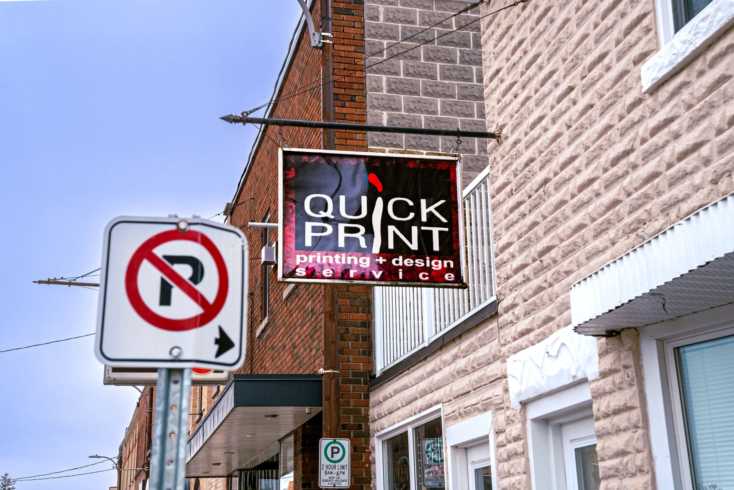Sign for Quick Print printing and design service hanging on a brick building with a patterned brick facade, a no parking sign and a parking meter nearby, under a clear sky.