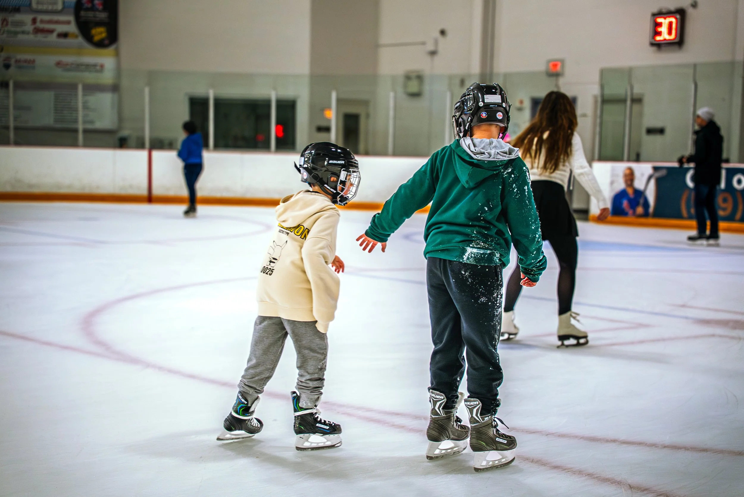 Two children wearing helmets and skates on an indoor ice rink, holding hands, with other skaters in the background.