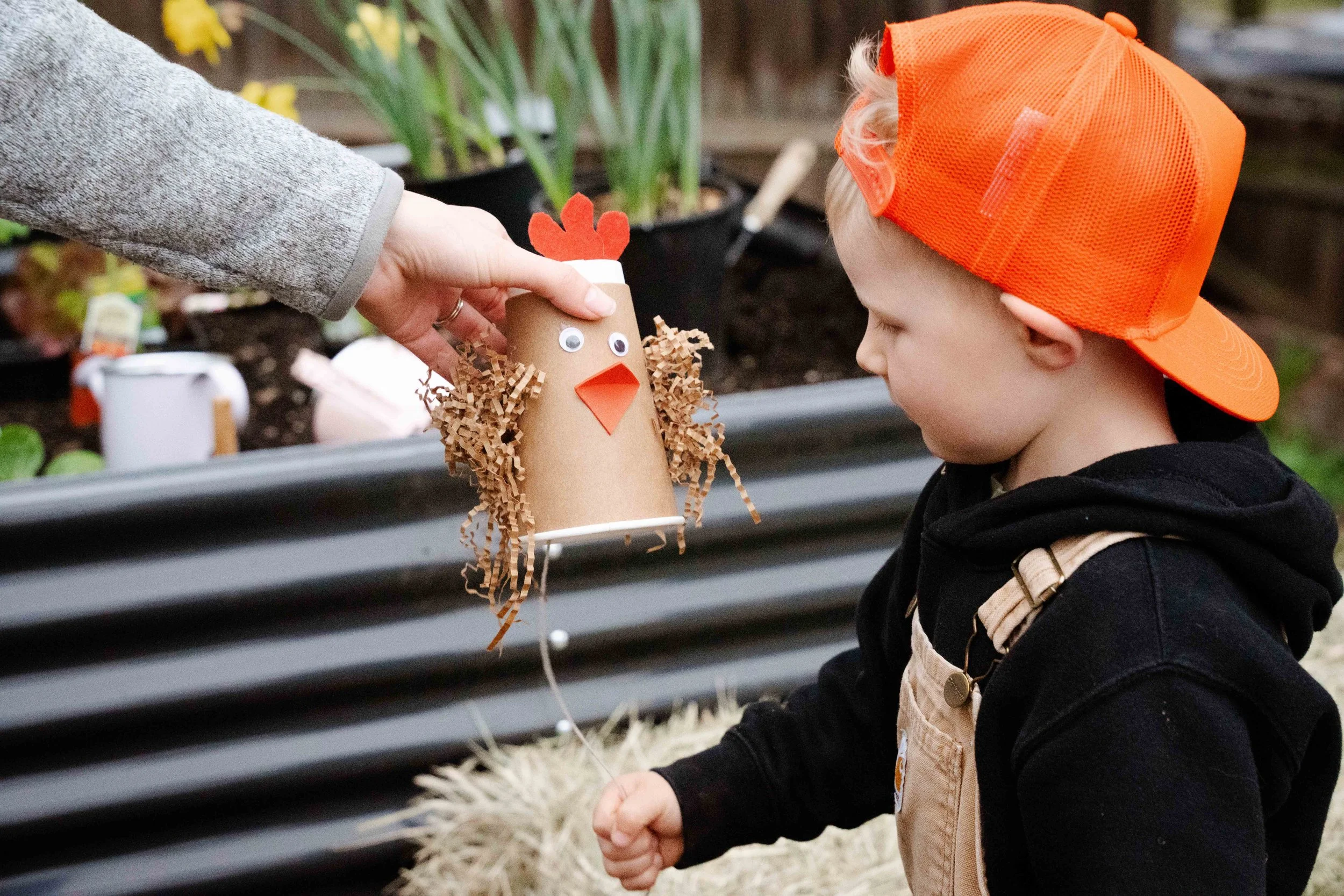 A child in an orange cap and beige overalls receiving a gift from an adult in a grey sweater. The gift is a craft paper turkey with googly eyes, a paper beak, and tissue paper feathers. They are outdoors near garden plants.
