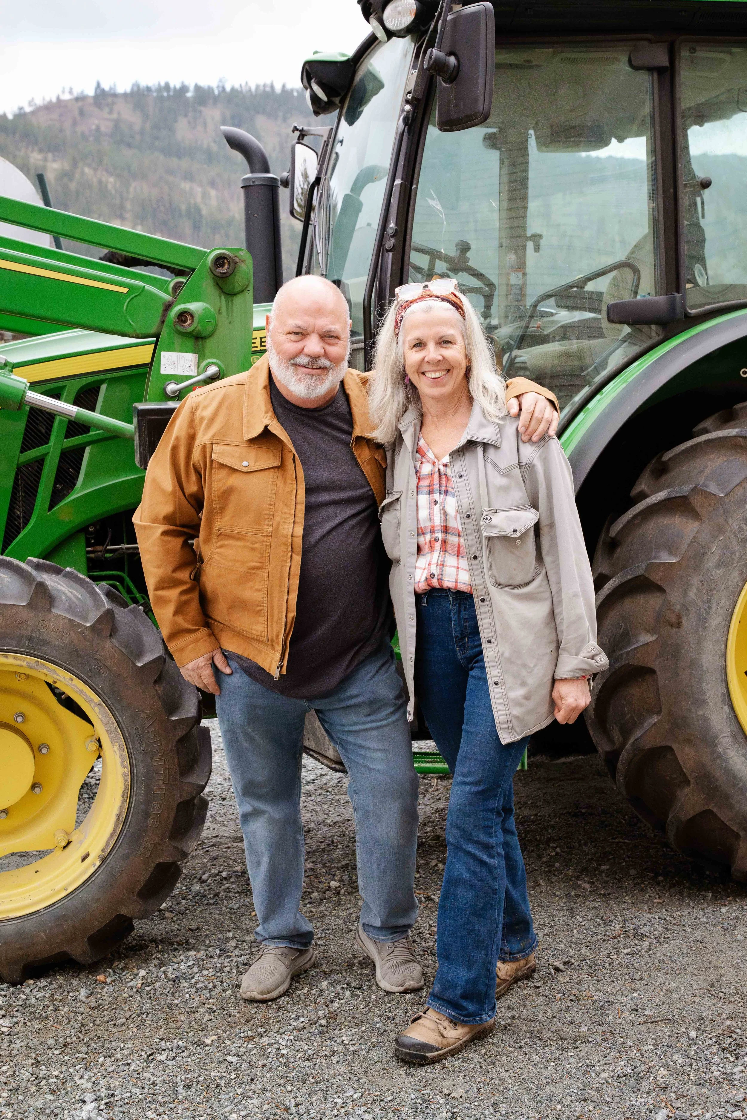 A smiling man and woman standing next to a green tractor outdoors on rocky ground with hills and trees in the background. The man is bald with a beard, wearing a brown jacket and jeans. The woman has long gray hair, sunglasses on her head, wearing a light gray jacket, plaid shirt, and jeans. They are close together, with the man’s arm around the woman.