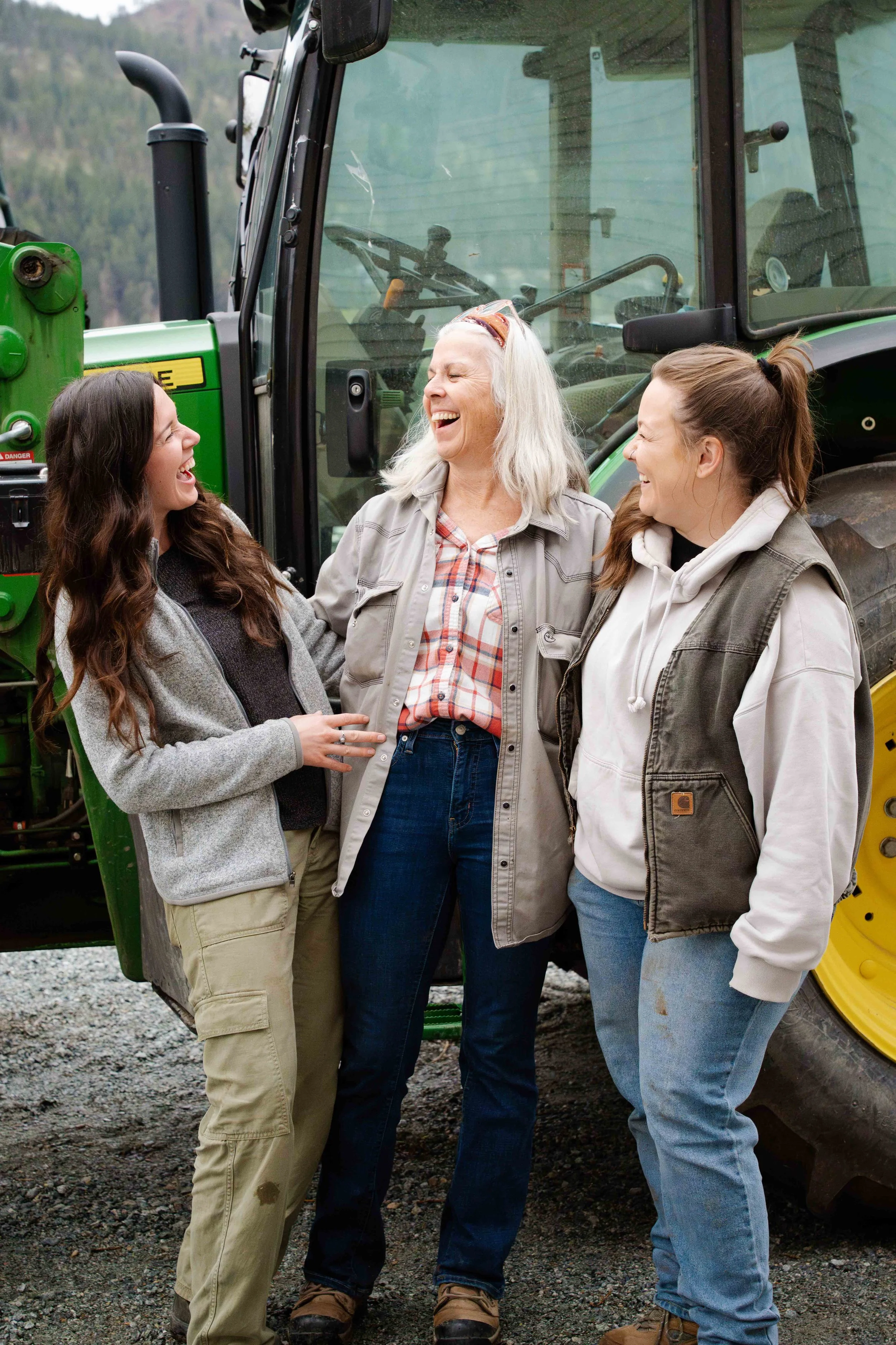 Three women standing in front of a green tractor, smiling and laughing together.