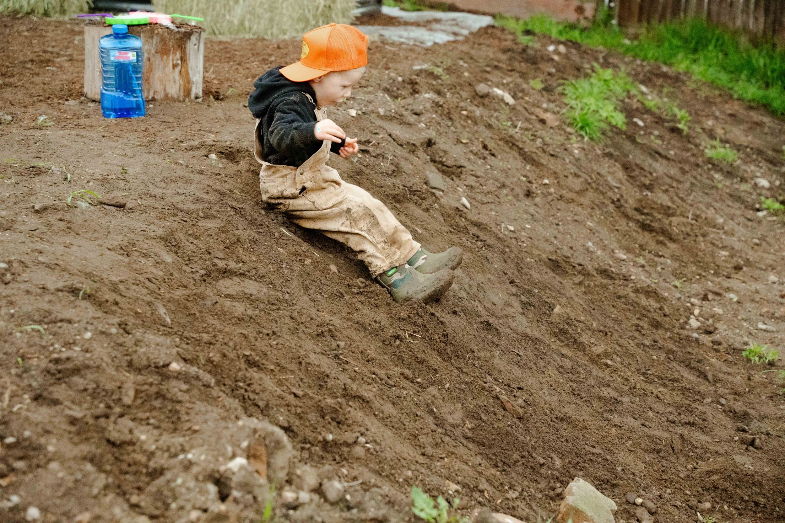 A young child wearing a black hoodie, tan overalls, green rain boots, and an orange cap sitting on a dirt slope outdoors, with a water bottle and a toy helicopter on a tree stump nearby.