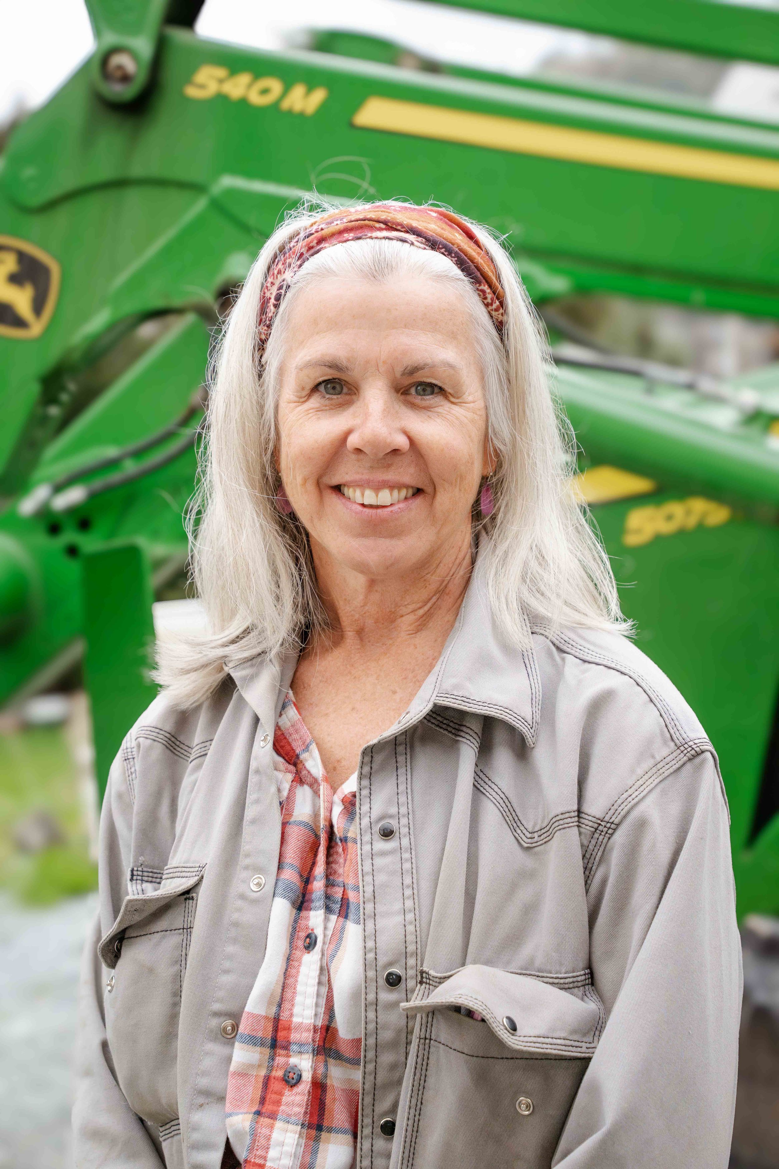 A smiling middle-aged woman with long gray hair, wearing a beige jacket over a plaid shirt, standing outdoors in front of a large green John Deere tractor.