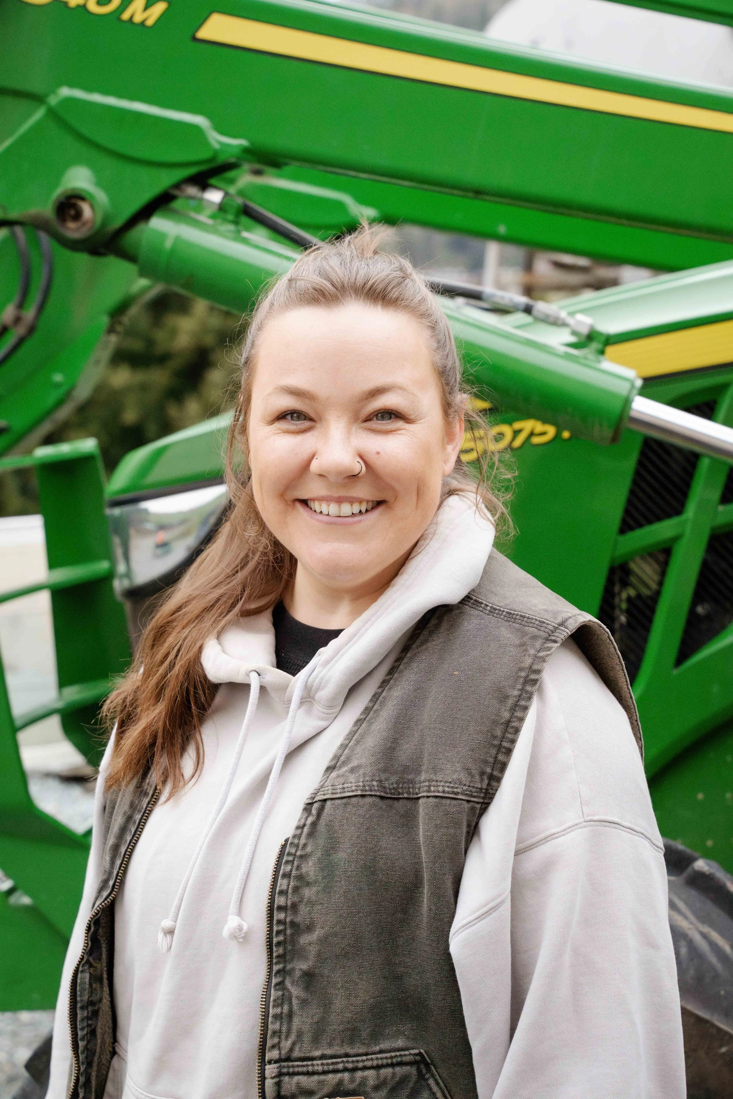 A young woman with long hair, smiling, wearing a white hoodie and a tan vest, standing outdoors in front of a green piece of heavy machinery.