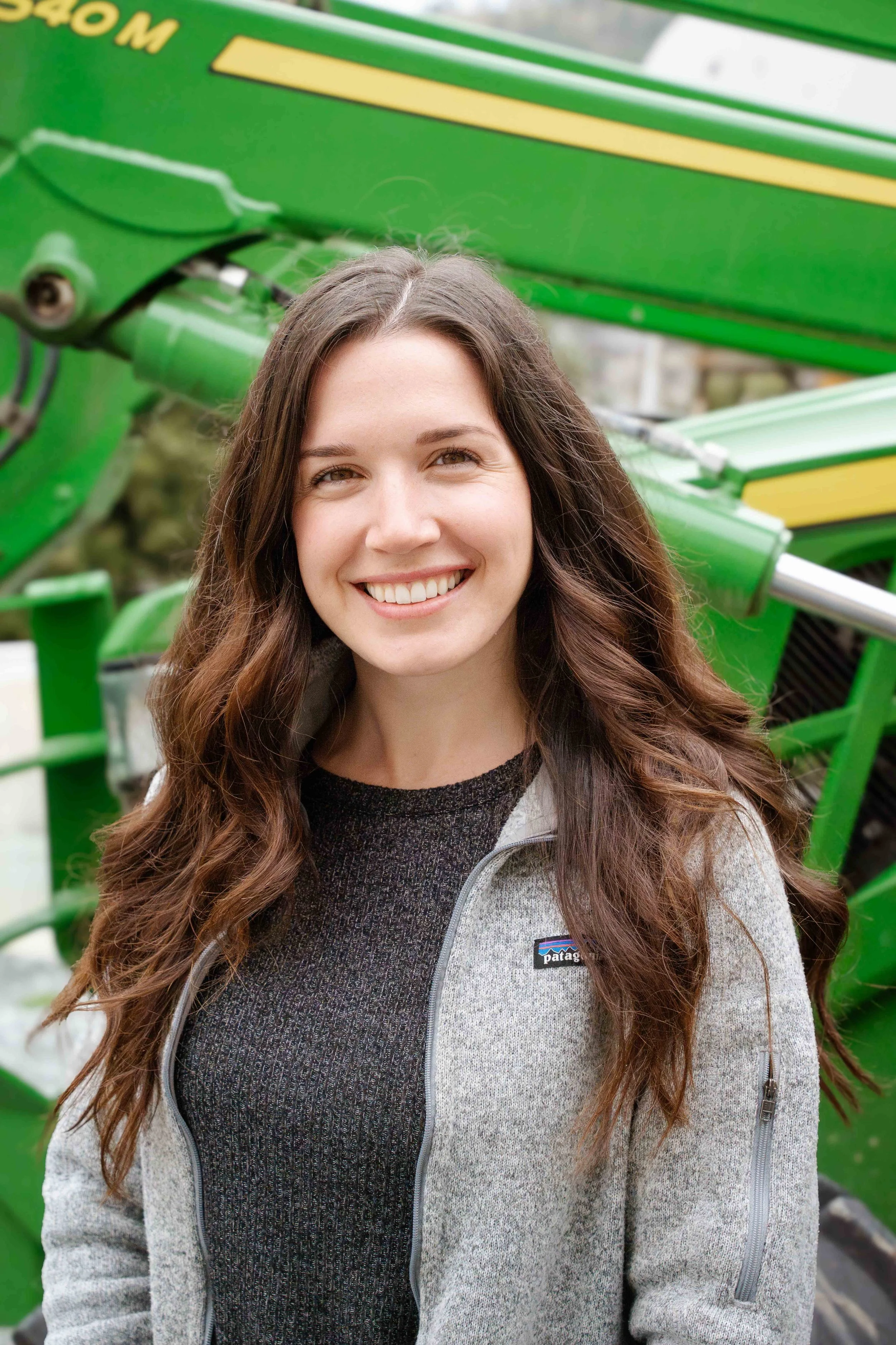 A woman with long brown hair smiling, wearing a gray Patagucci jacket and a dark top, standing outdoors near a green piece of machinery.