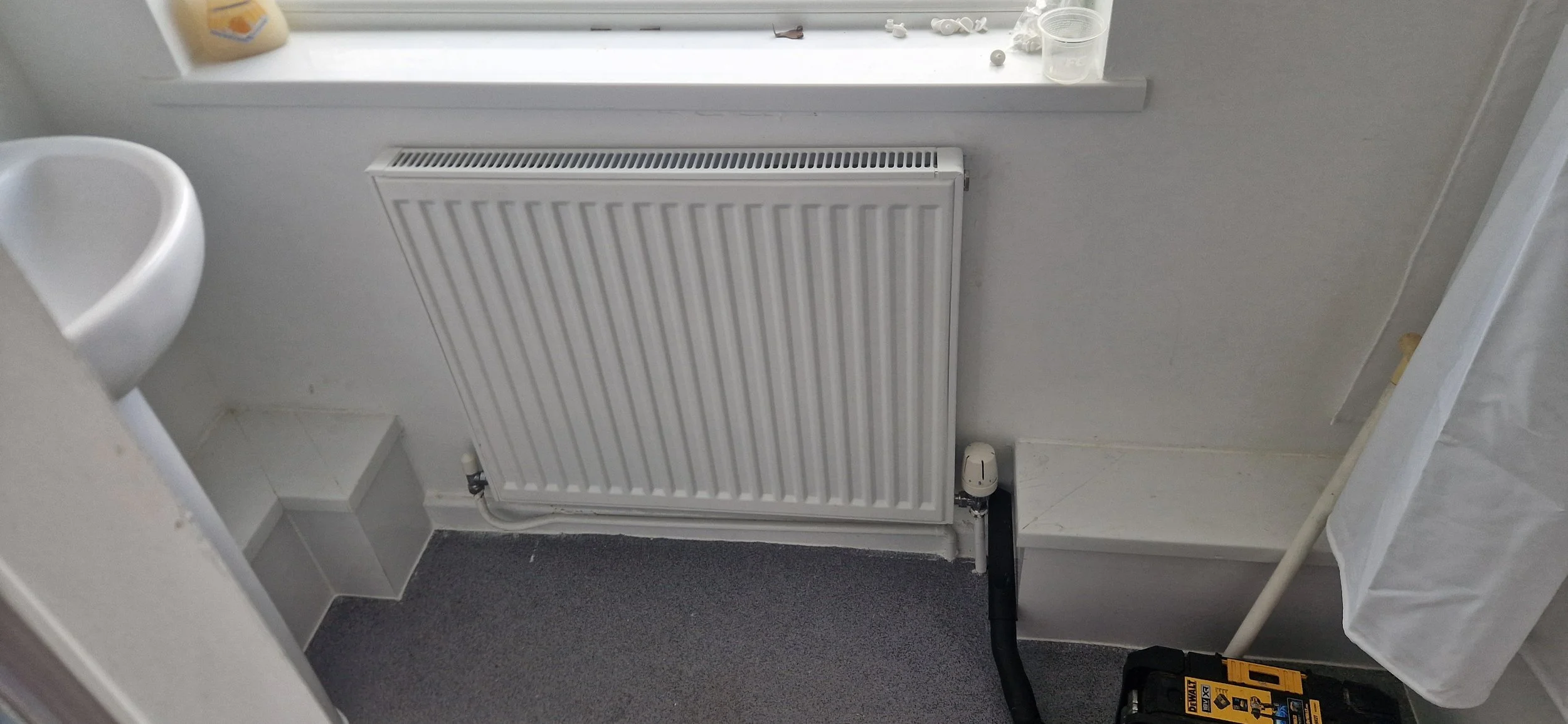 A radiator installed on a white wall near a window with a windowsill, with a few small objects on the window ledge, next to a white toilet and a gray carpeted floor.