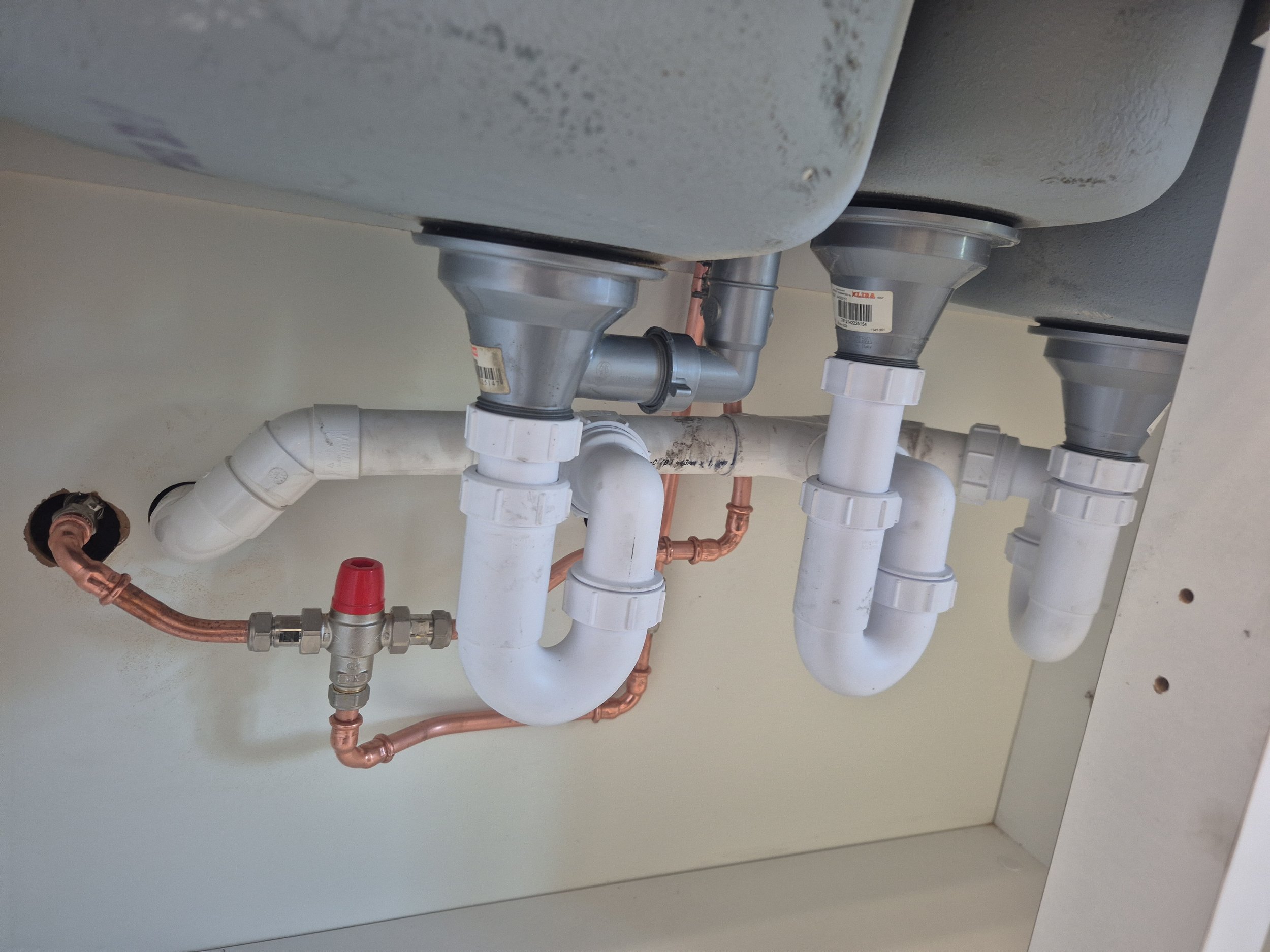 Plumbing pipes and fittings beneath a sink, including copper and PVC pipes, with some labels visible.