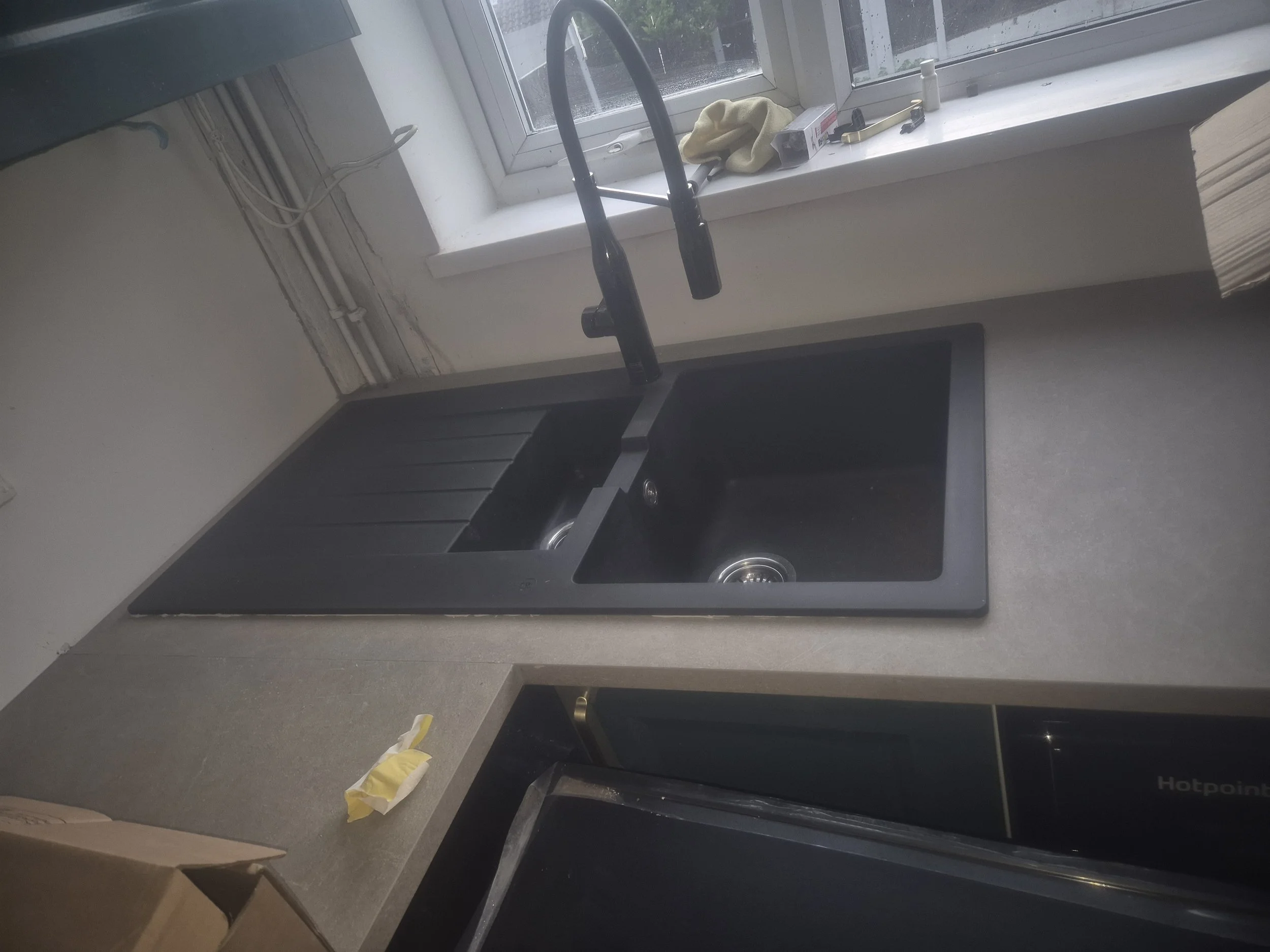 A kitchen sink area with a black granite countertop, a black sink, and a window above it. Items on the windowsill include a yellow cloth, a white bottle, a box, and tools. There is a yellow glove on the counter near the cabinet door.