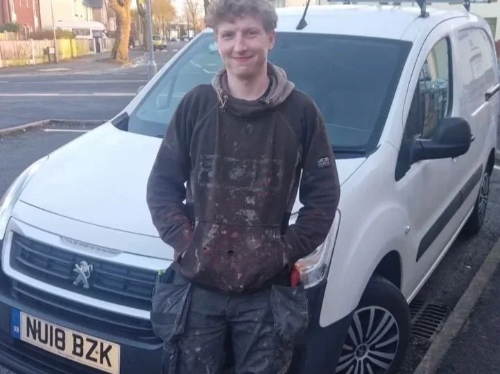 Young man in work clothes standing in front of a white van in an outdoor parking lot, smiling at the camera.