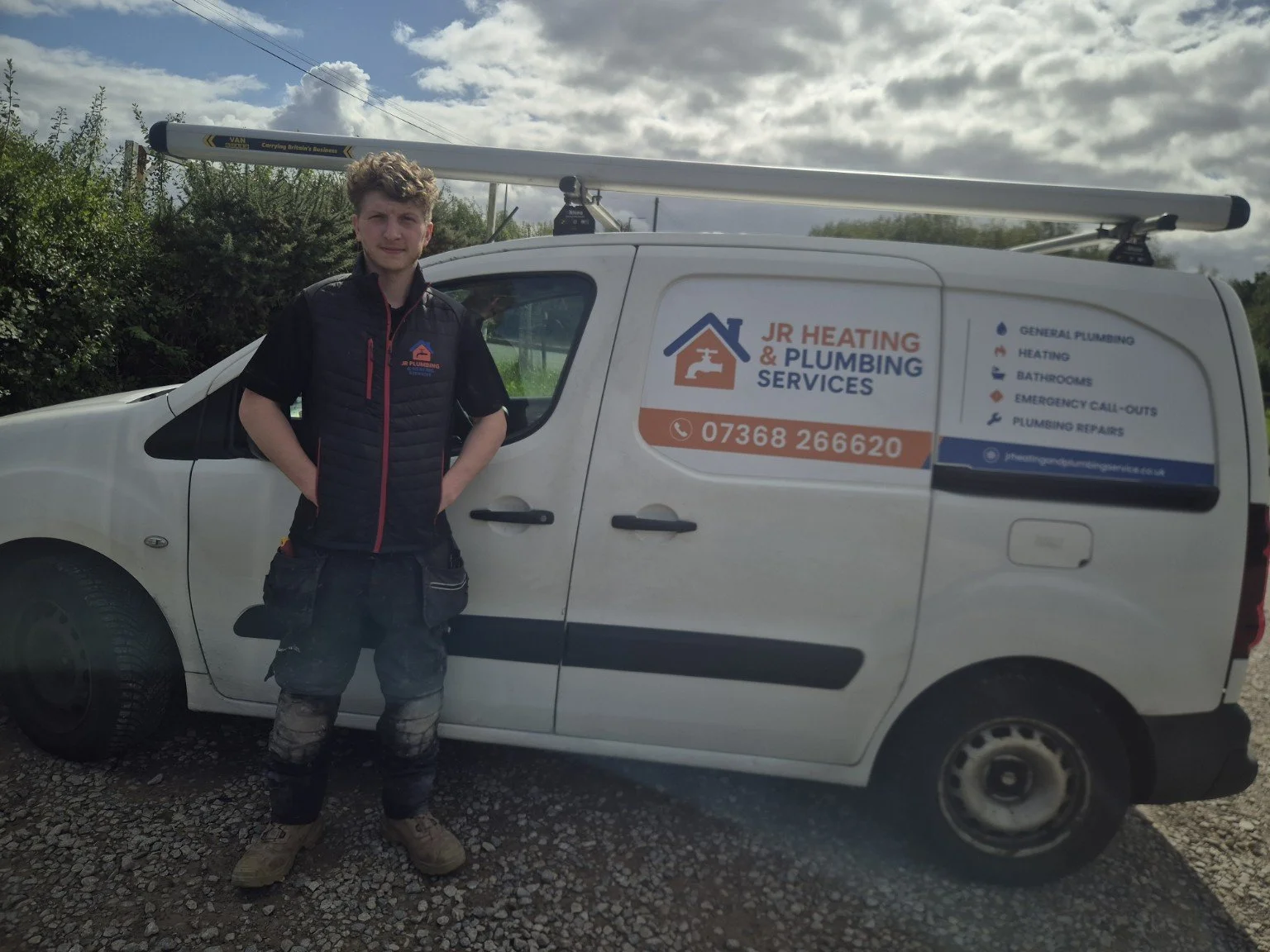 Young man in branded work clothes standing beside a white JR Heating & Plumbing Services van outdoors, smiling at the camera.