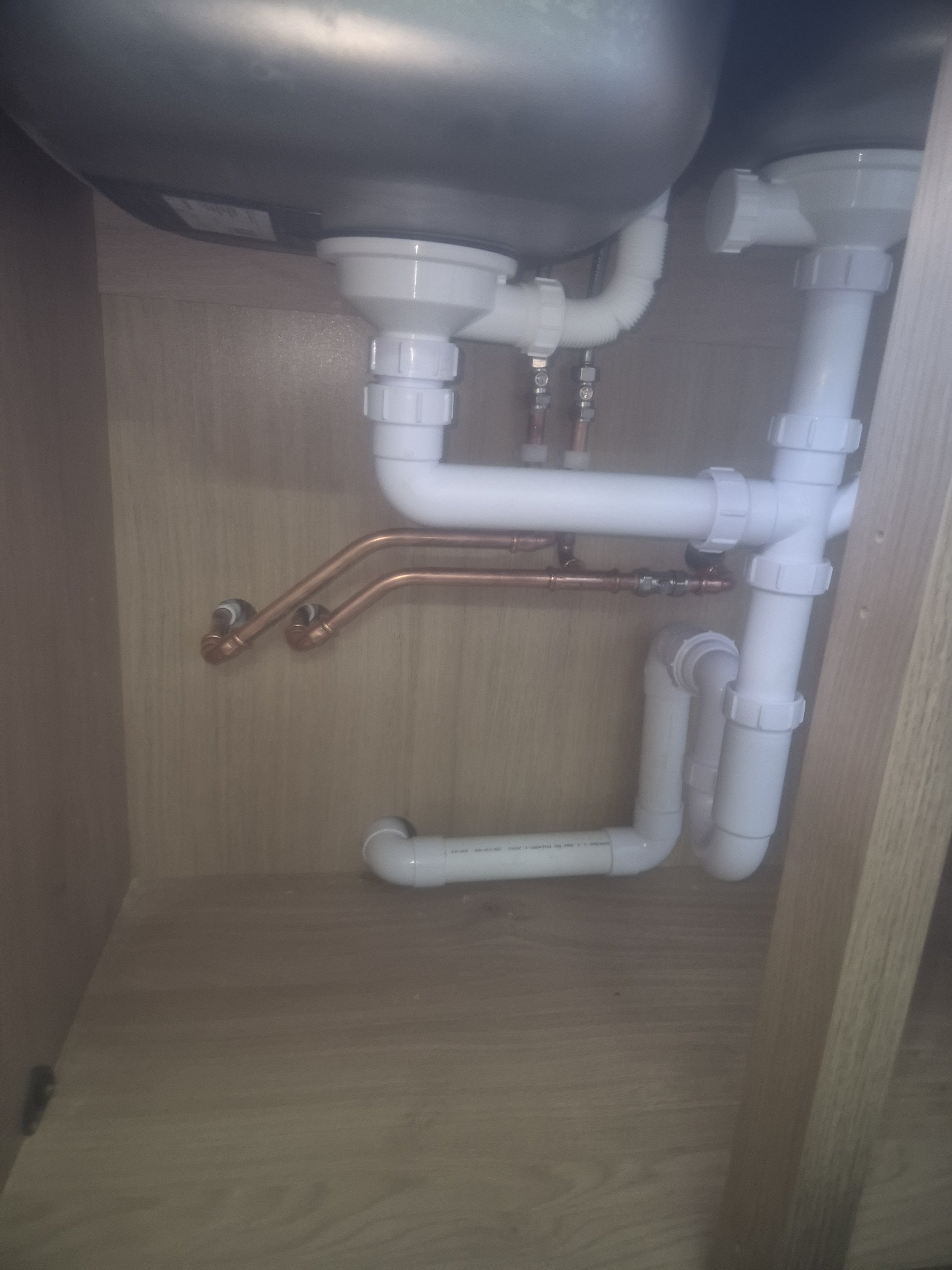 Underneath a kitchen sink showing white PVC pipes and copper pipes connected to plumbing fixtures.