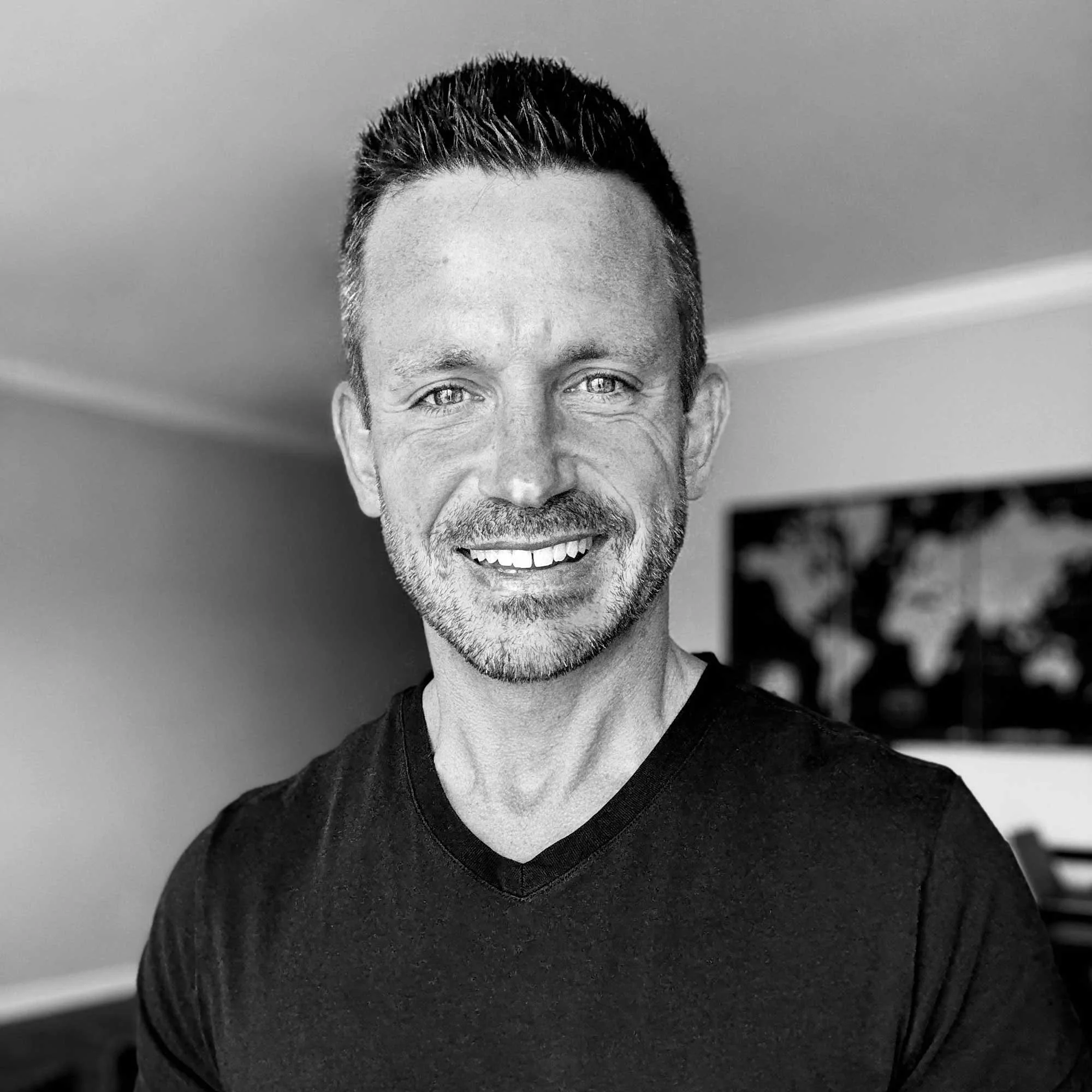 A smiling man with short hair and a short beard wearing a black V-neck shirt in an indoor setting.