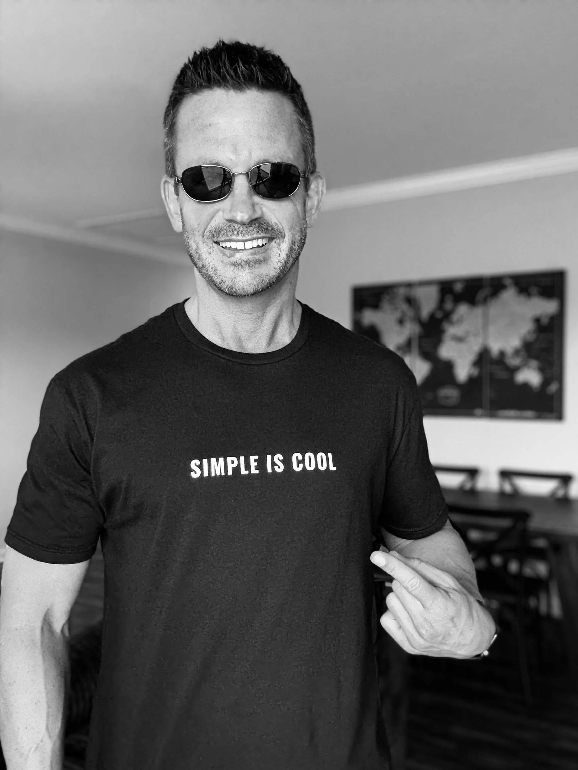 A man with short dark hair, sunglasses, and a beard is smiling, wearing a black T-shirt that says "SIMPLE IS COOL", and is pointing to the text with his right hand, in a room with a world map on the wall behind him.