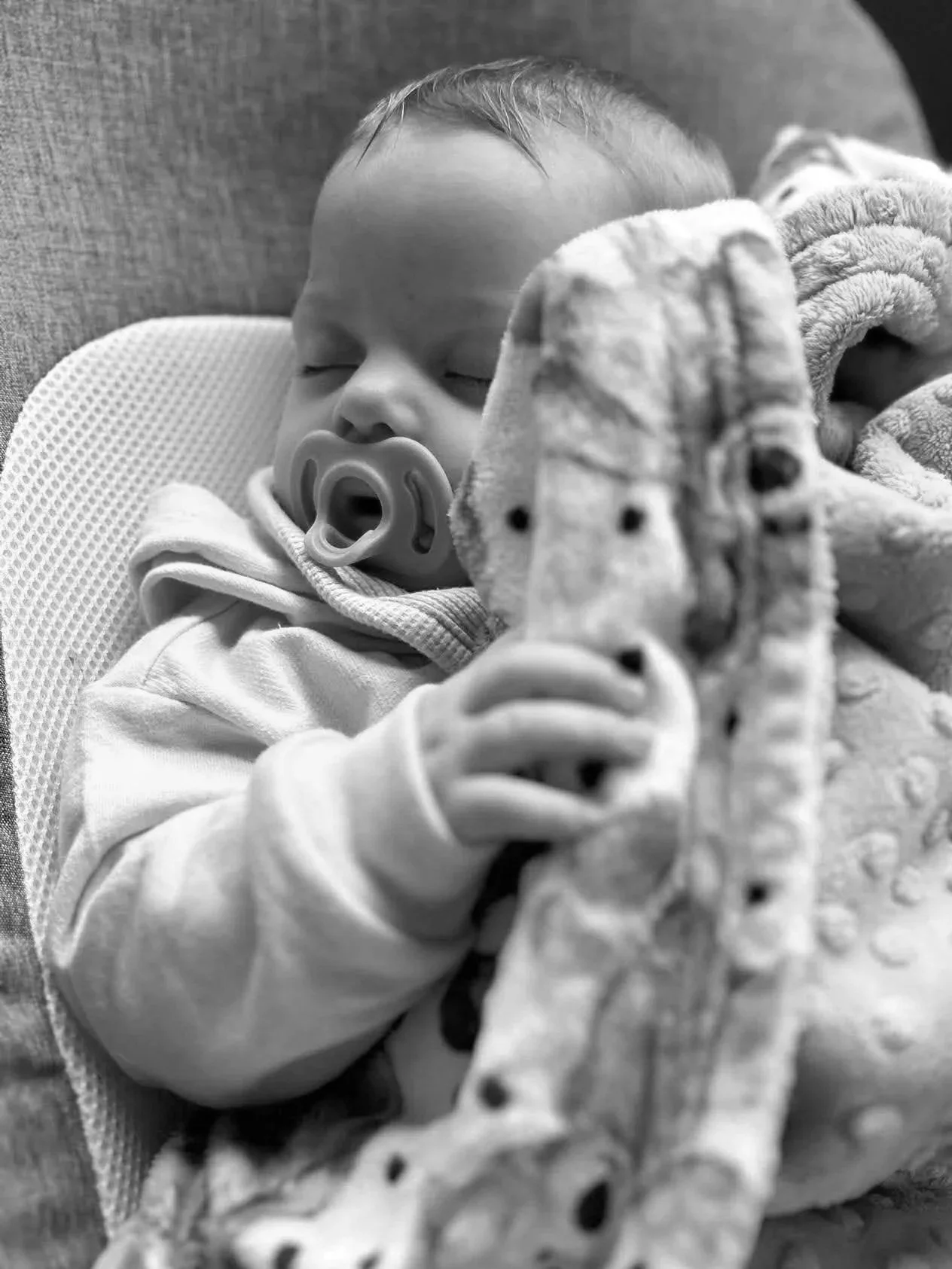 Black and white photo of a sleeping baby wearing a pacifier, lying on a textured surface, with a fuzzy blanket partially covering the face.