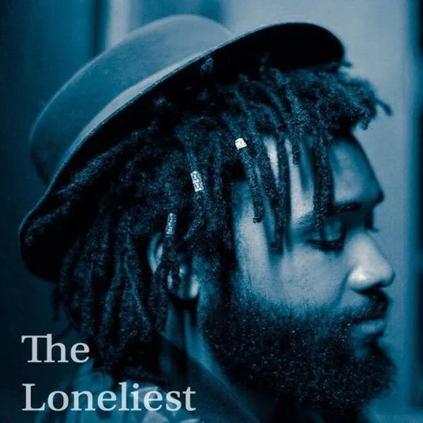 Close-up of a man with dreadlocks, wearing a hat, in a blue-toned portrait. Text reads: "The Loneliest".