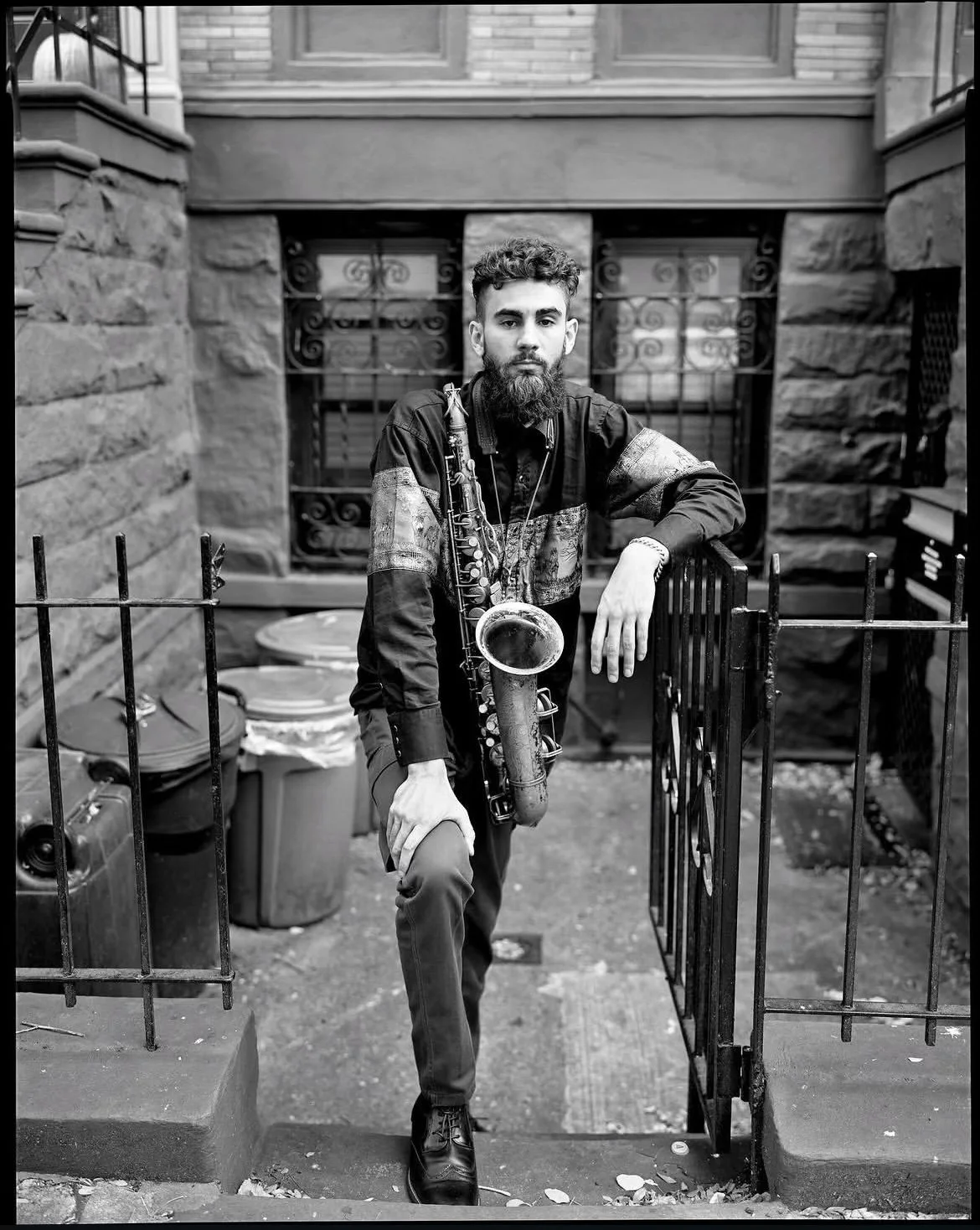 A man with a beard wearing a patterned shirt standing in a doorway, holding a saxophone, outside in an urban alley with trash bins and stone walls.