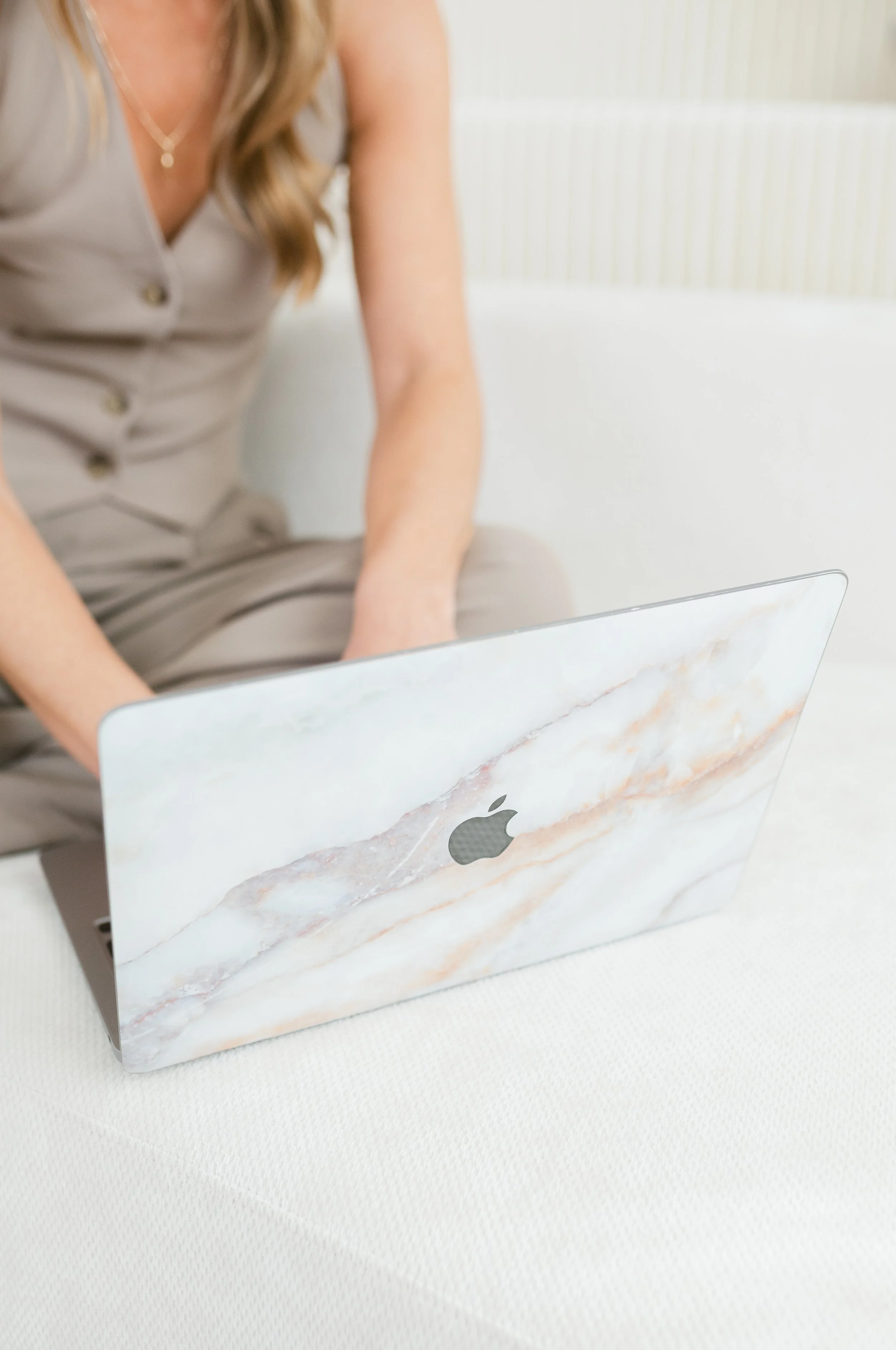 Close-up of a woman wearing beige dress using a marble-patterned MacBook laptop, on a white textured surface.