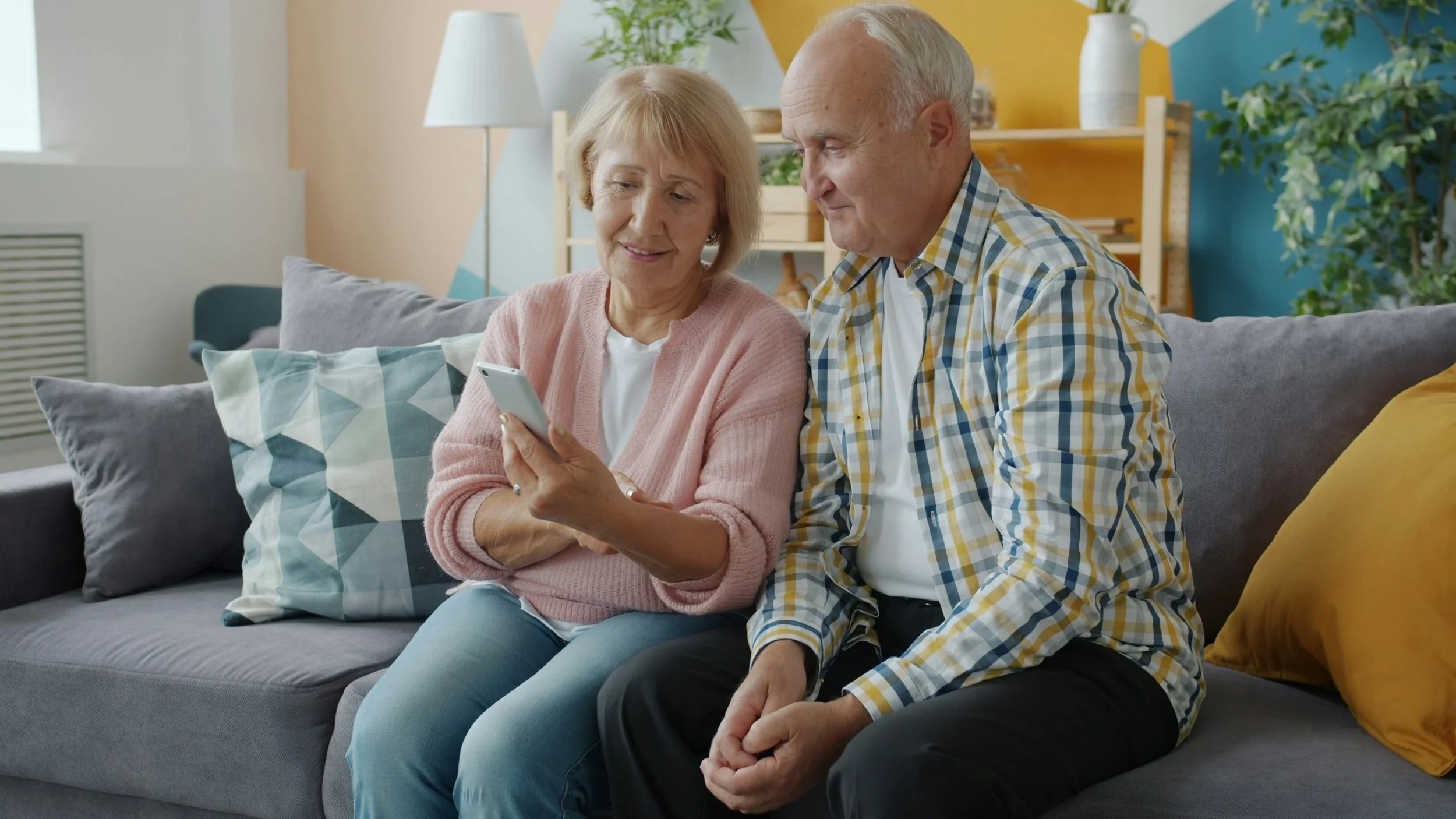An elderly woman and man sitting on a gray couch, looking at a smartphone together in a brightly decorated living room.