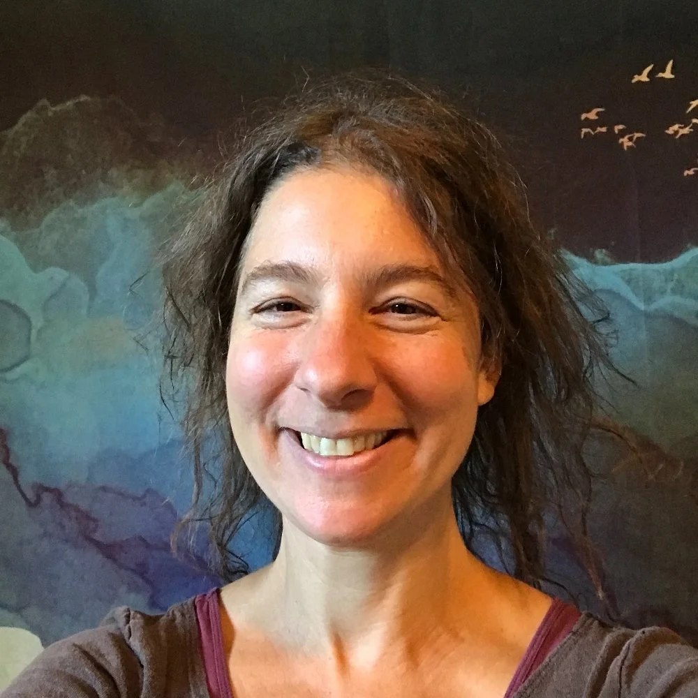 A woman with curly hair smiling in front of a dark world map mural with birds flying.