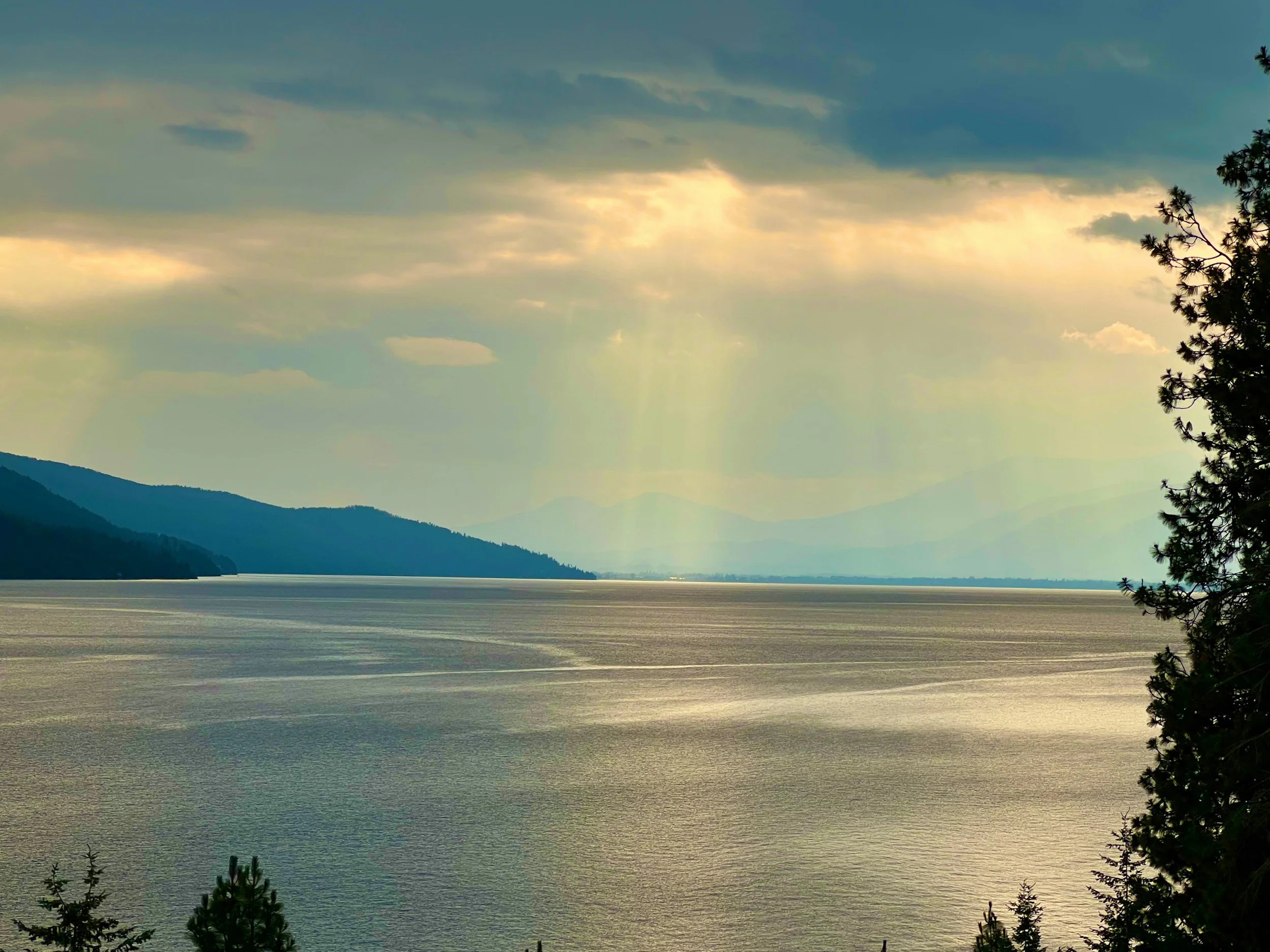 Scenic landscape of a large lake with mountains in the distance, partly cloudy sky with sunlight breaking through.